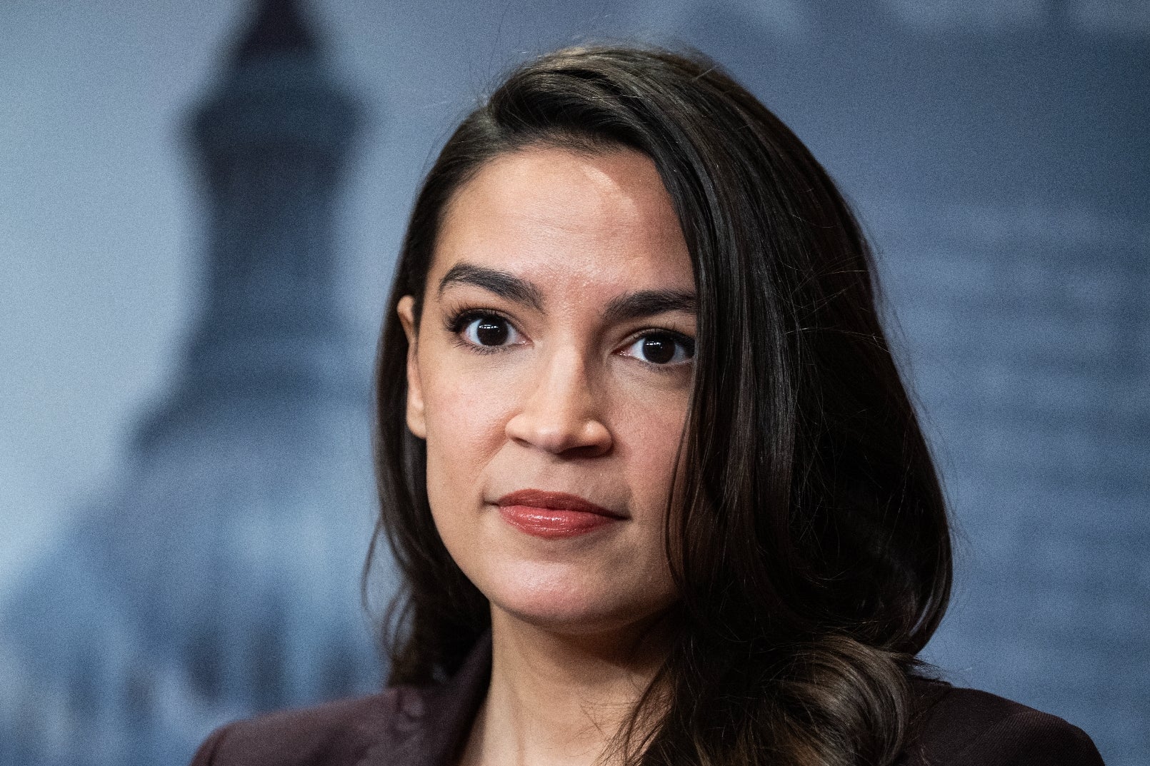 A person in formal attire stands in front of a blurred cityscape background with the U.S. Capitol dome visible