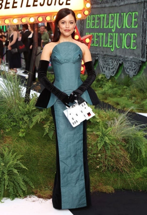 Person successful a strapless gown with agelong gloves, holding a themed clutch, connected a reddish carpet astatine an lawsuit with "Beetlejuice" signage