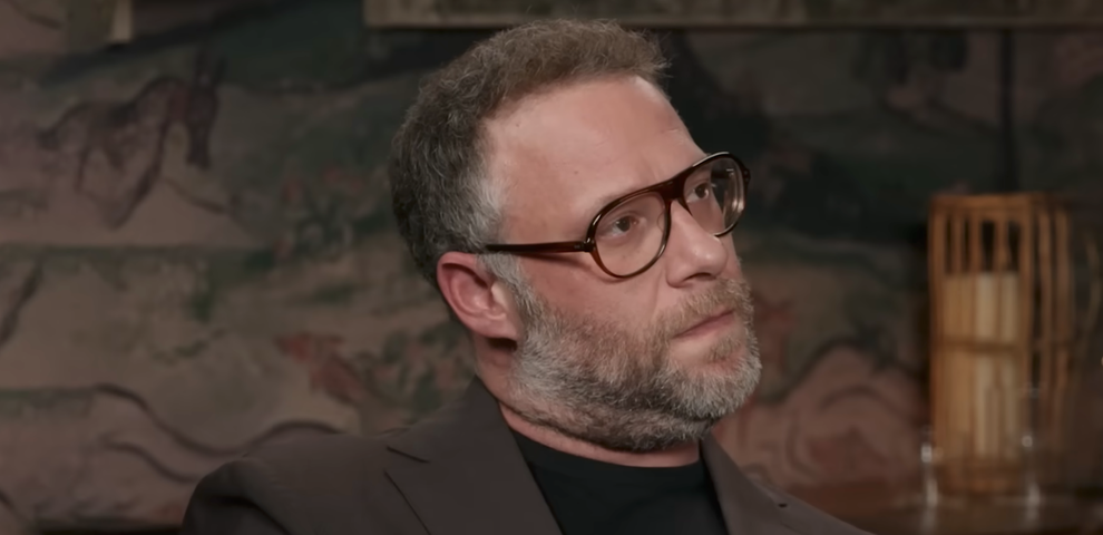 A person with glasses and a beard looks to the side, wearing a suit jacket