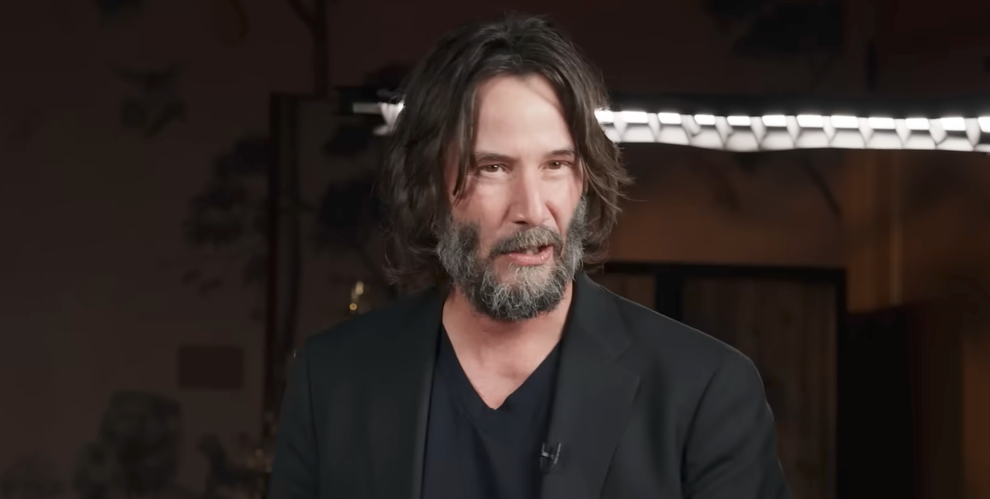 A person with long hair and a beard is wearing a black jacket over a T-shirt, speaking in front of studio lighting