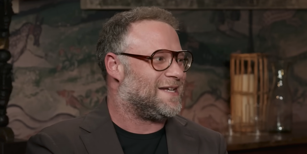 Man with glasses and beard speaking in an indoor setting, wearing a blazer over a t-shirt