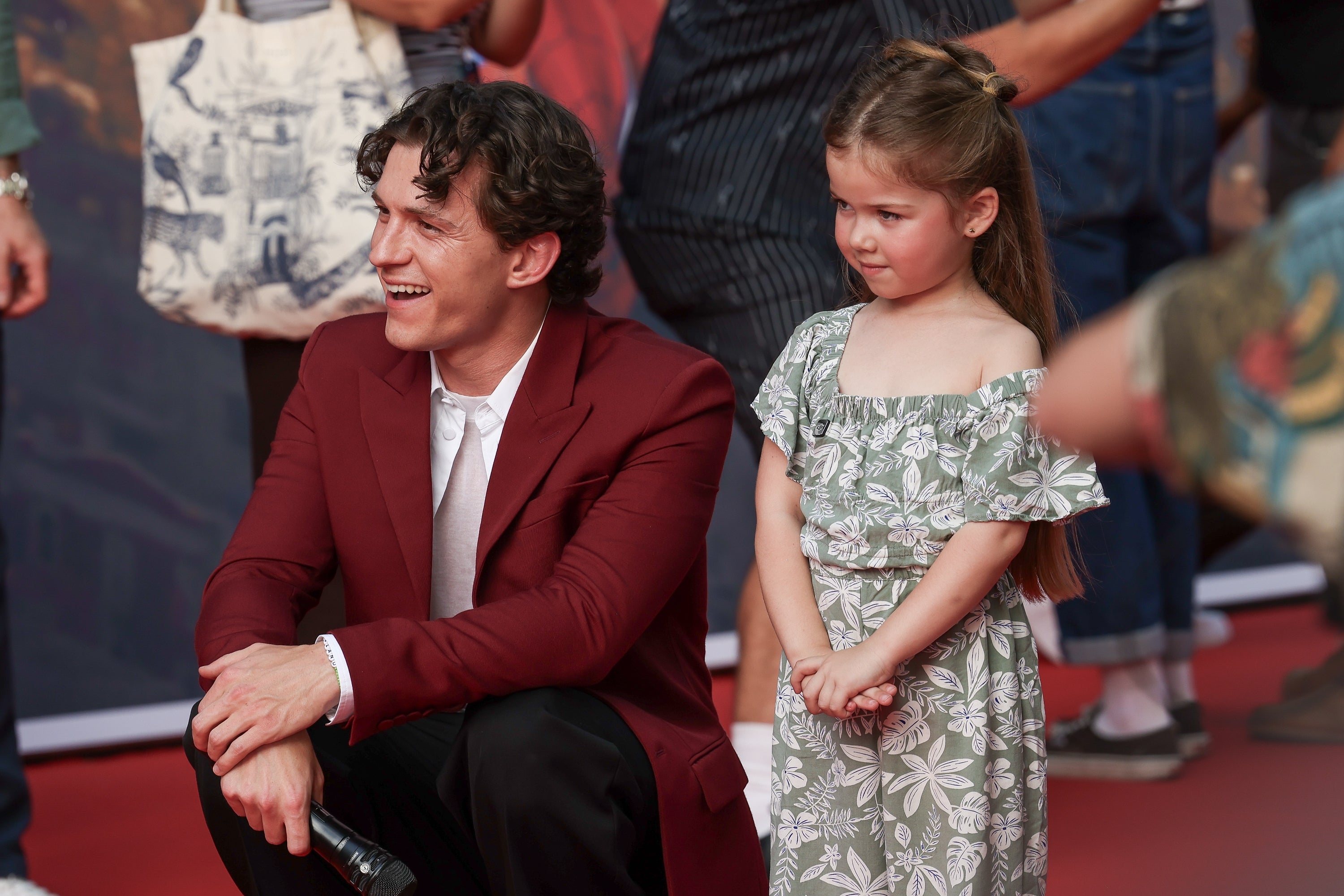 Man successful a stylish suit kneels connected a reddish carpet beside a young miss successful a floral dress, some smiling pleasantly astatine an event