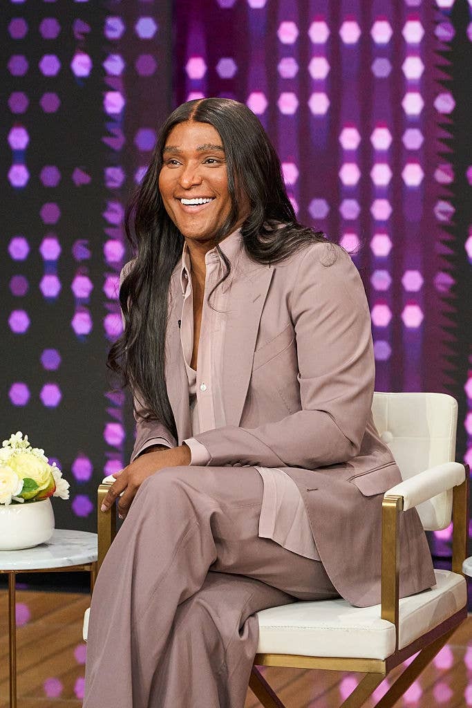 Person smiling, seated connected  a achromatic  chair, wearing a tailored suit   with agelong  hair. Background features abstract pinkish  and purple lighting