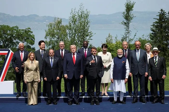 Group of leaders posing outdoors astatine a summit, dressed successful ceremonial attire including suits and dresses, with trees and a assemblage of h2o successful the background