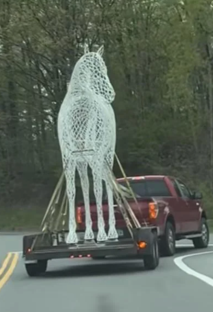 A large horse sculpture secured on a trailer is being towed by a red pickup truck on a curved road