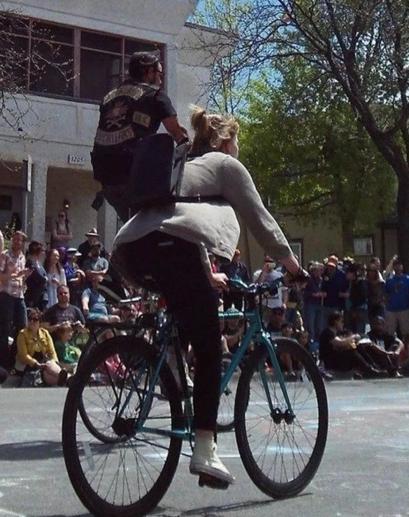 A woman rides a bicycle with a man sitting on the back, balancing in an amusing position, surrounded by a crowd at an outdoor event