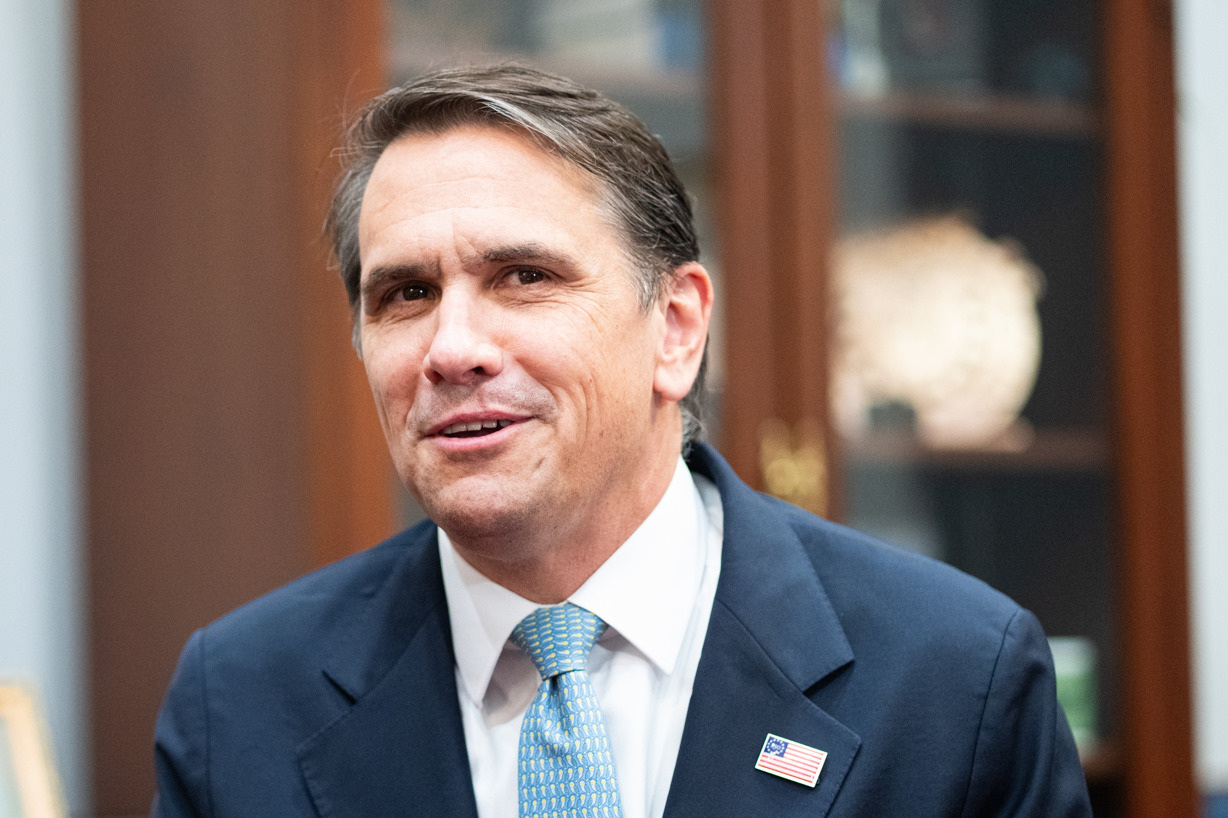 A idiosyncratic   successful  a suit   with a tiny  emblem  pin connected  the lapel, smiling, lasting  indoors with a blurred background