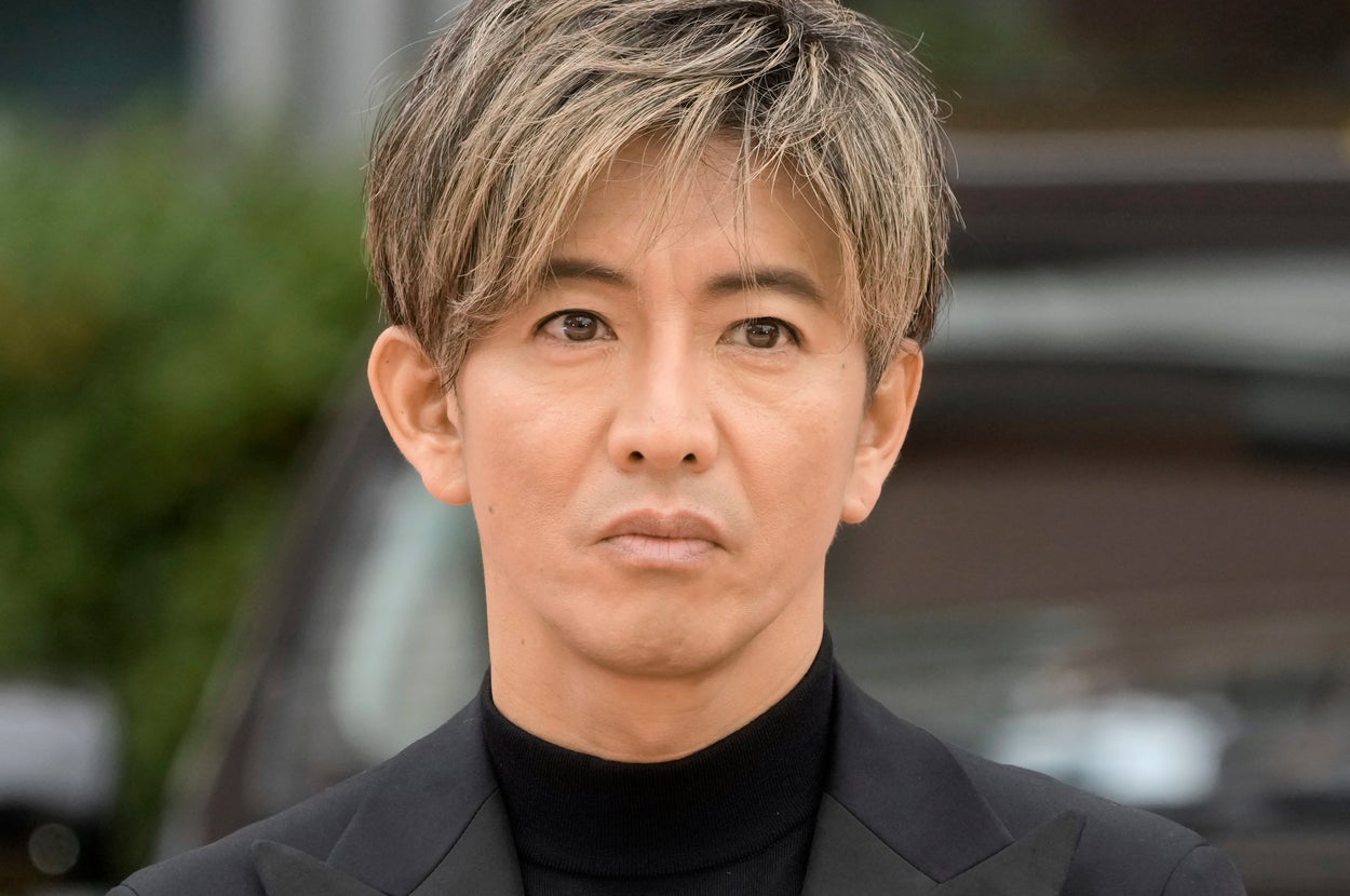著名人が黒のタートルネックとジャケットを着てカメラを見つめている。背景にタクシーの一部が見える。