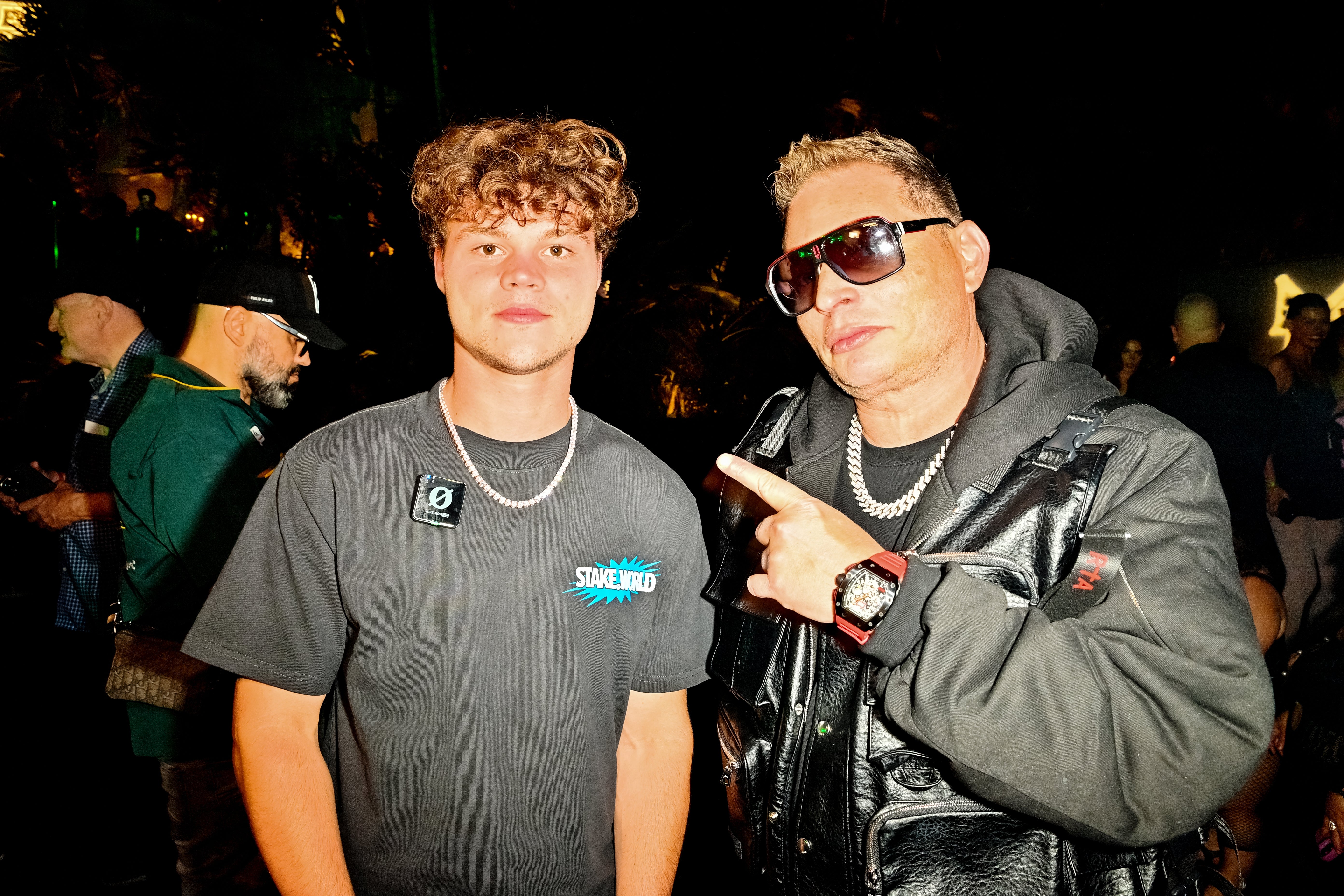 Jack Doherty and Scott Storch in a nighttime event; one in a casual tee, the other in a leather jacket and sunglasses, pointing at the first person