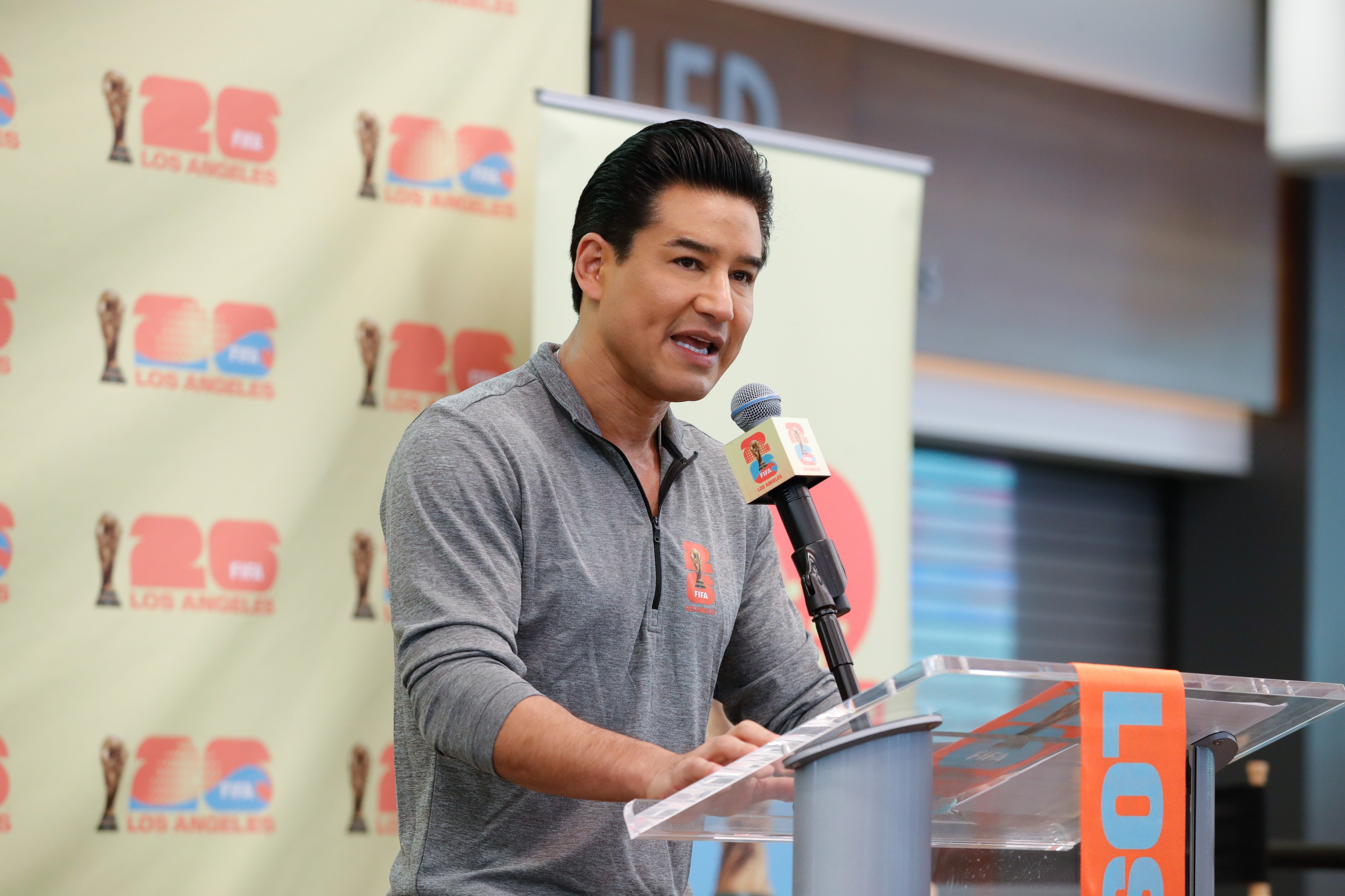 Mario Lopez speaking astatine a podium with Los Angeles 2026 backdrop, wearing a casual pullover, microphone successful hand
