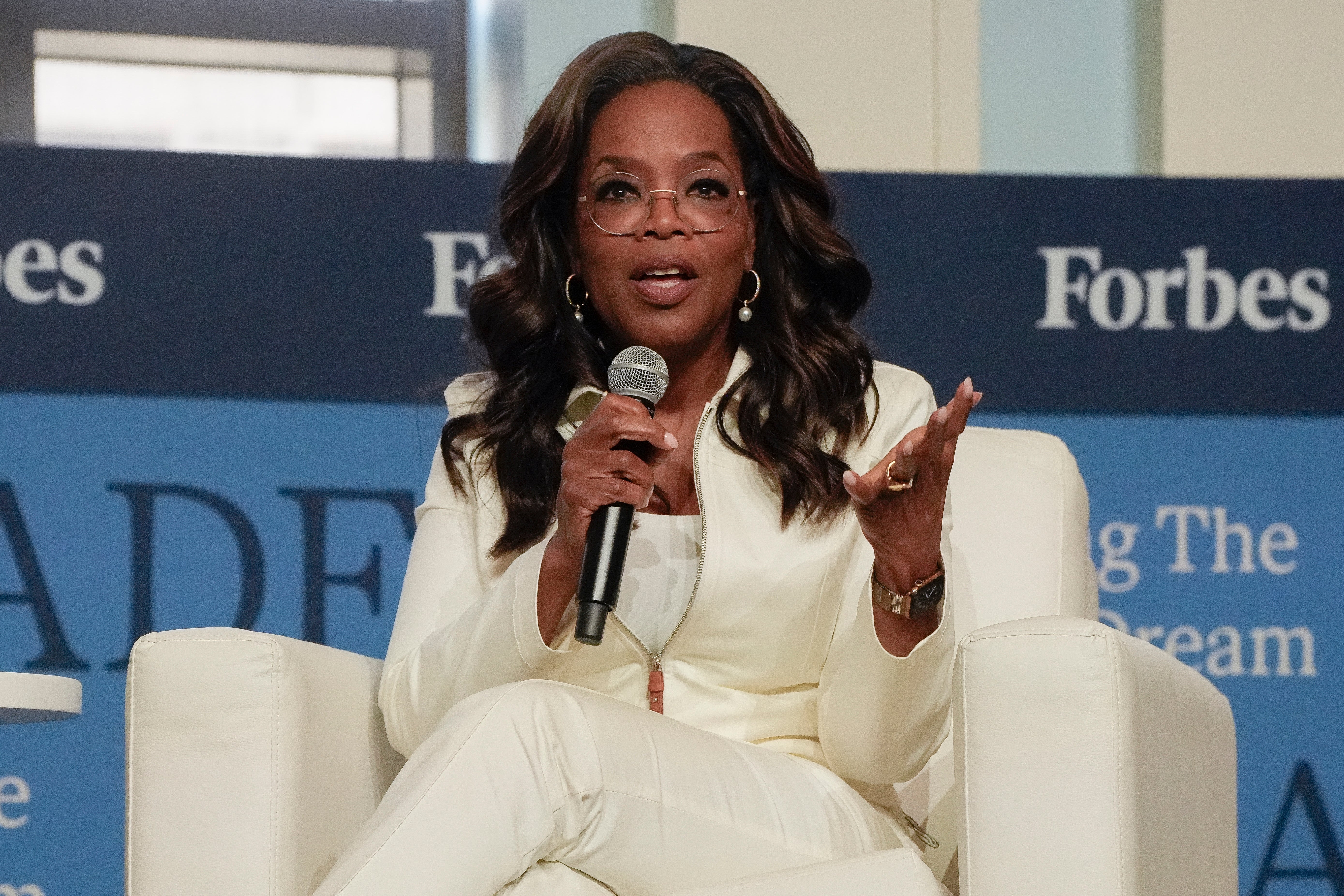 Oprah Winfrey in a white suit speaks into a microphone while sitting on stage at a Forbes event