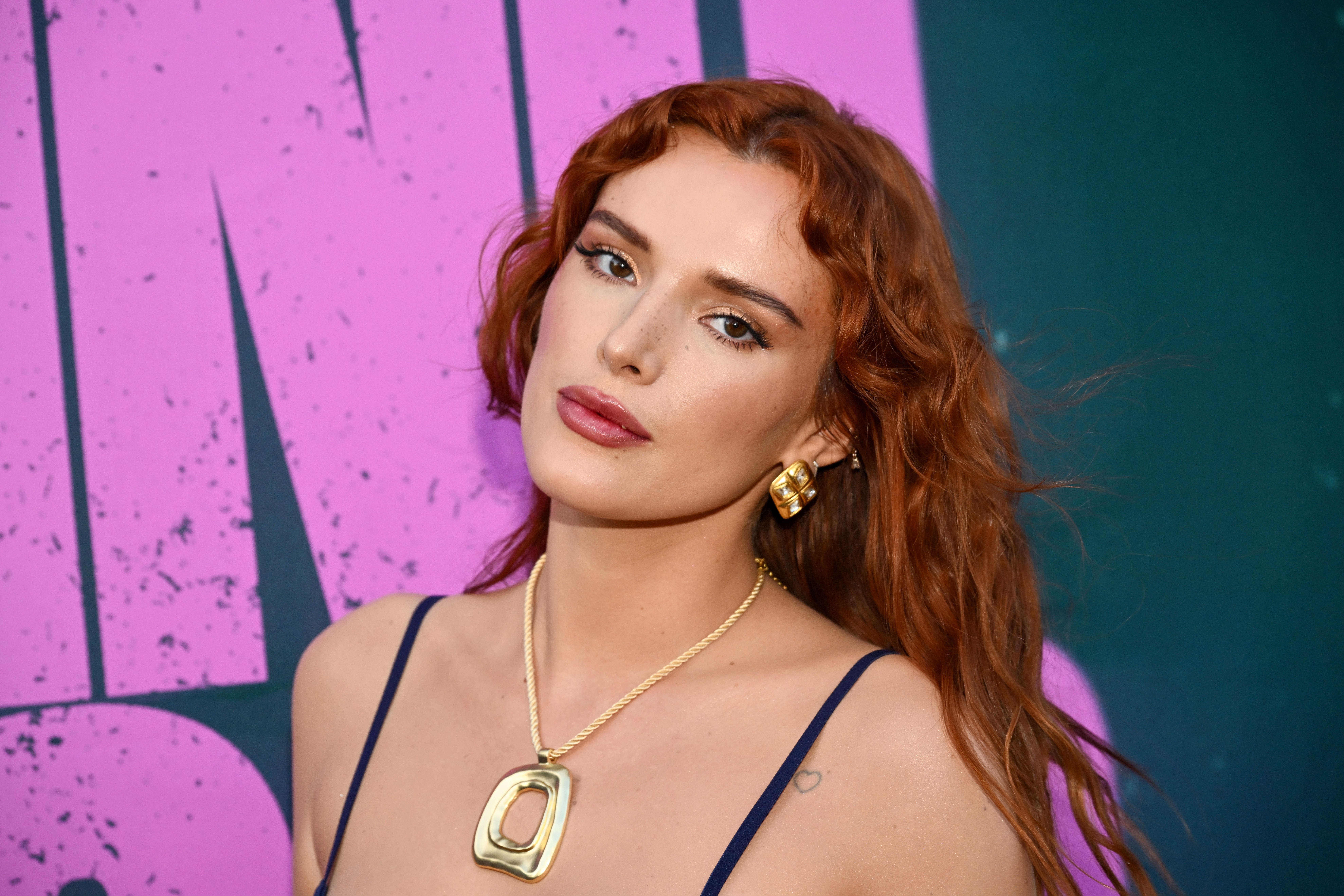 Bella Thorne posing on the red carpet, wearing a low-cut dress, gold jewelry, and wavy hair at a formal event