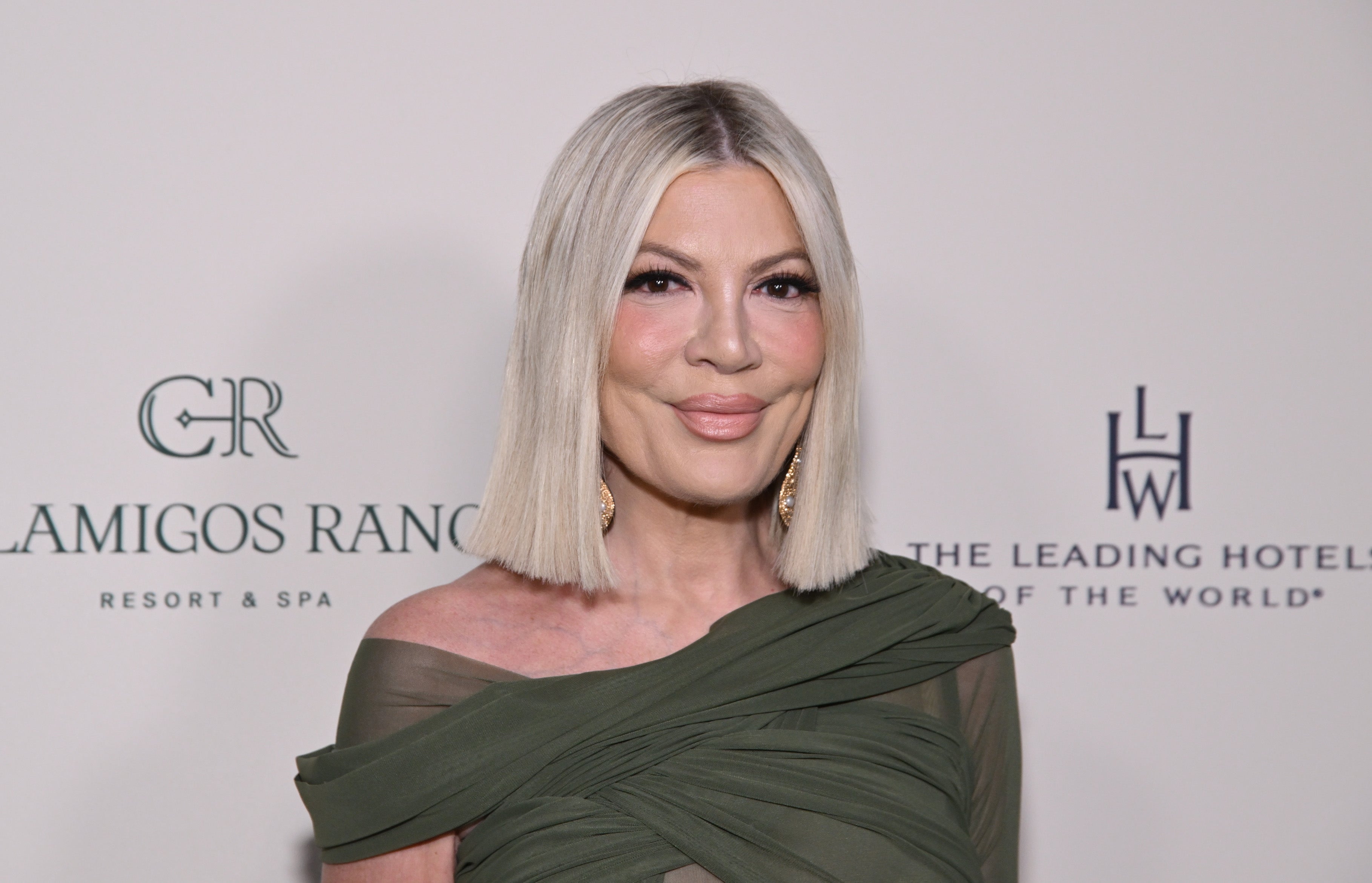 Tori Spelling with shoulder-length hair in off-shoulder, draped gown on event backdrop