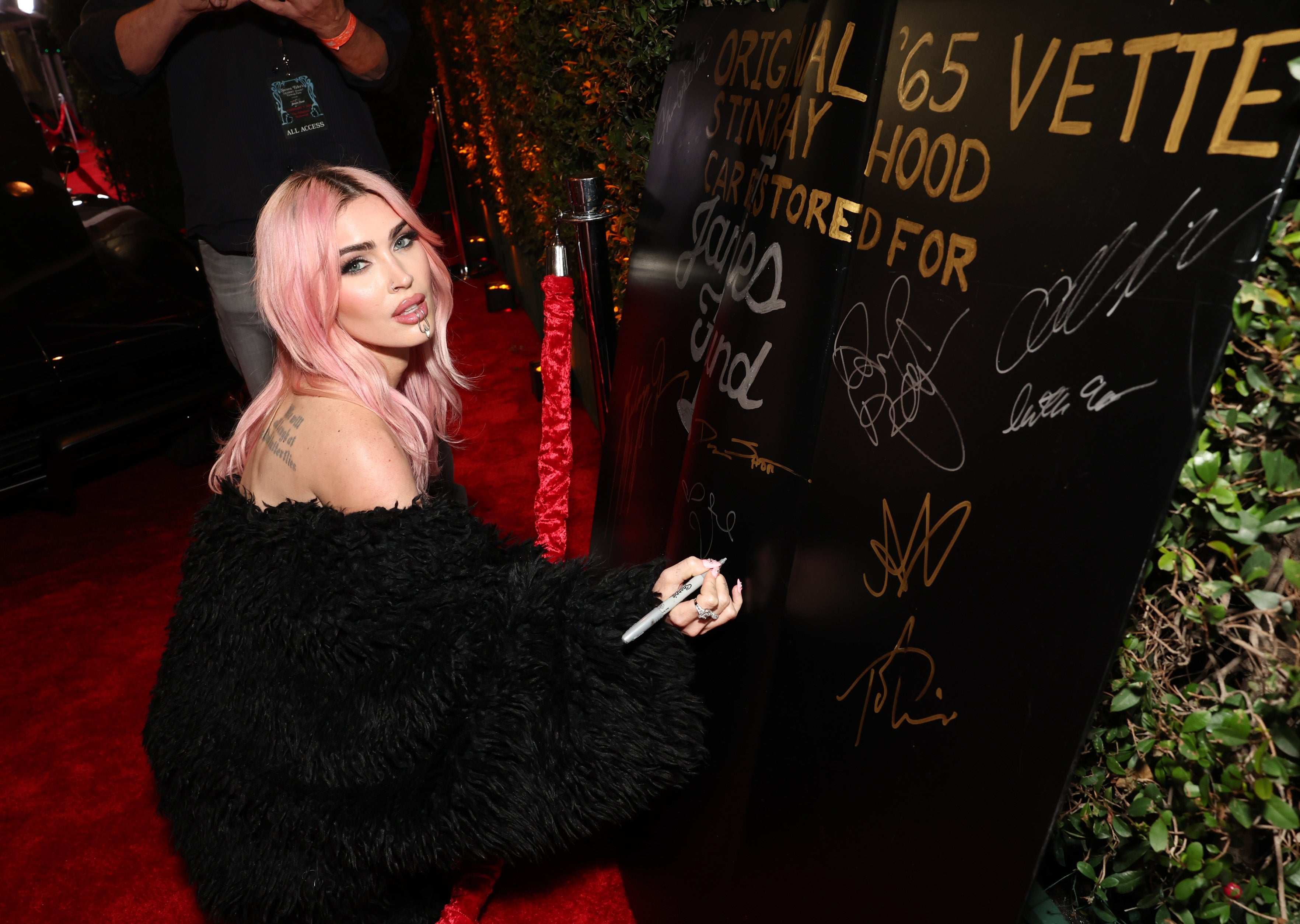Megan Fox with pink hair and a fur-like jacket signs a large autograph board at an event