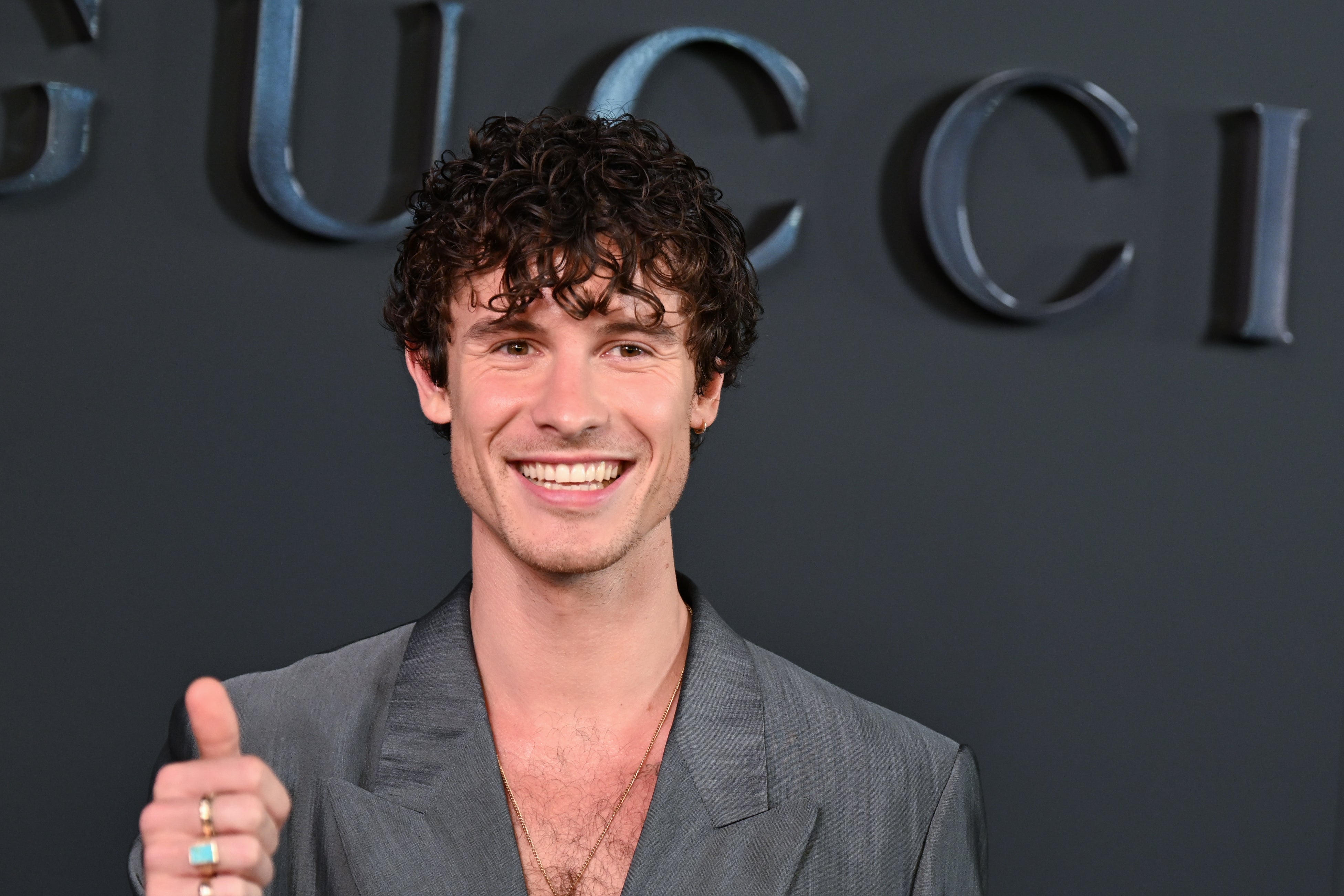 Shawn Mendes on red carpet in gray suit, smiling and giving thumbs up in front of a branded wall