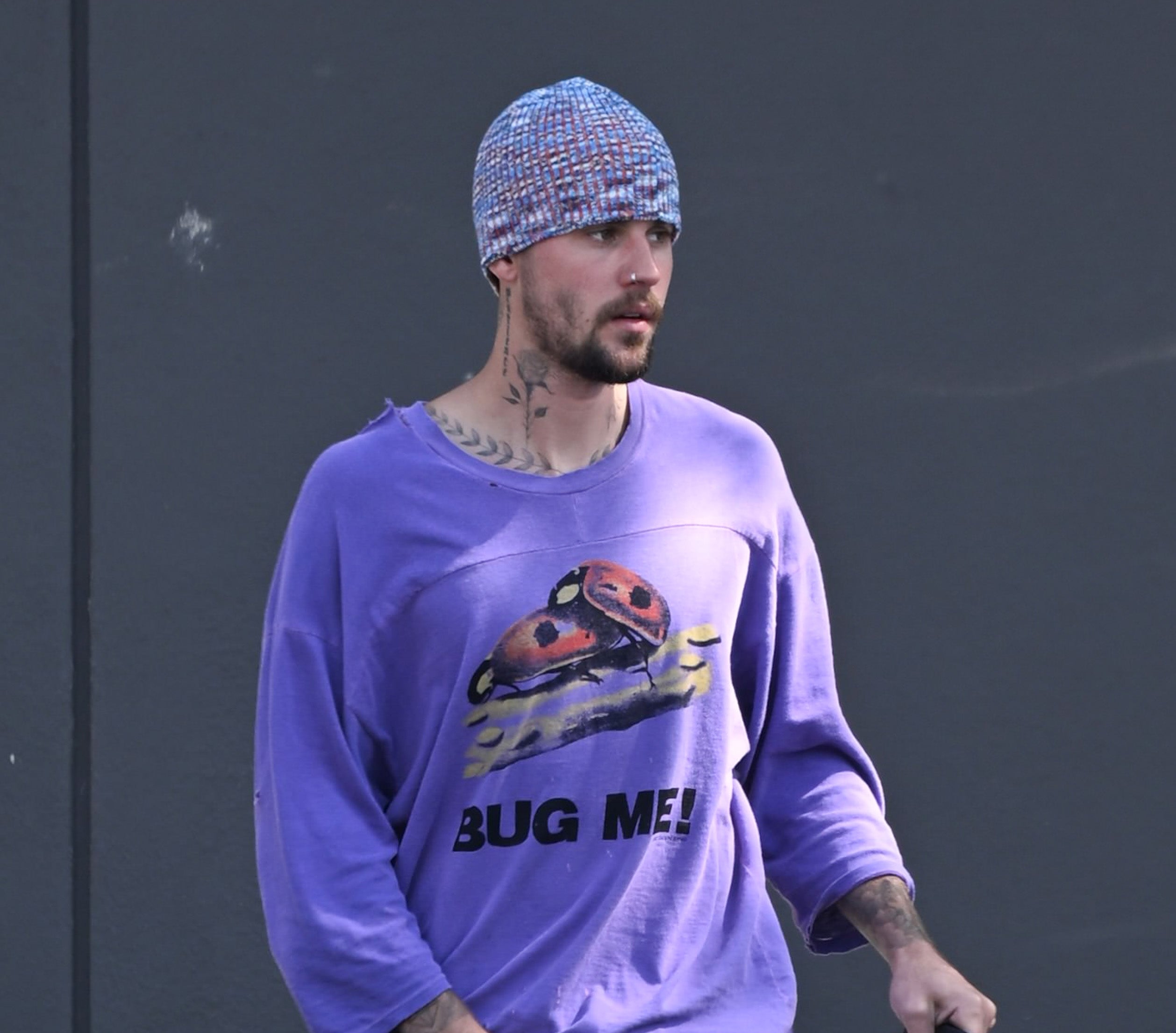 Justin Bieber wearing a beanie and a loose sweatshirt with a "Bug Me!" graphic and ladybug design, walking outside against a gray wall