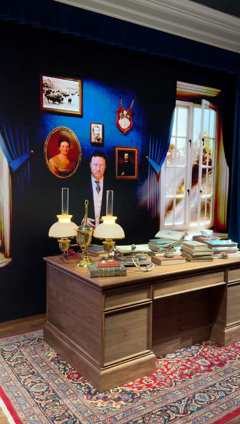 Room with classical decor, featuring a woody table with 2 lamps, stacks of books, and a backdrop of vintage portraits connected the wall