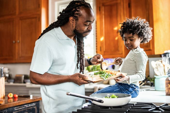 Pria dan anak-anak dengan gembira memasak bersama di dapur; pria itu sedang memotong bumbu sementara anak itu menambahkan bumbu ke dalam wajan