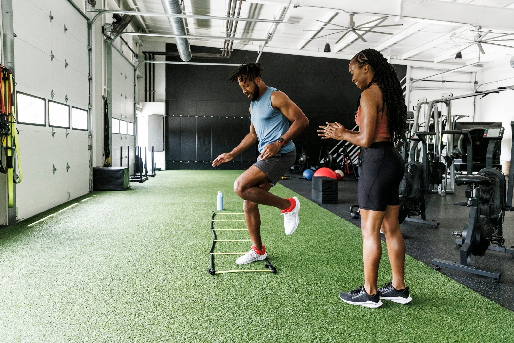 Dua orang berlatih di gym; yang satu melakukan latihan tangga sementara yang lain mengamati dan memberi instruksi