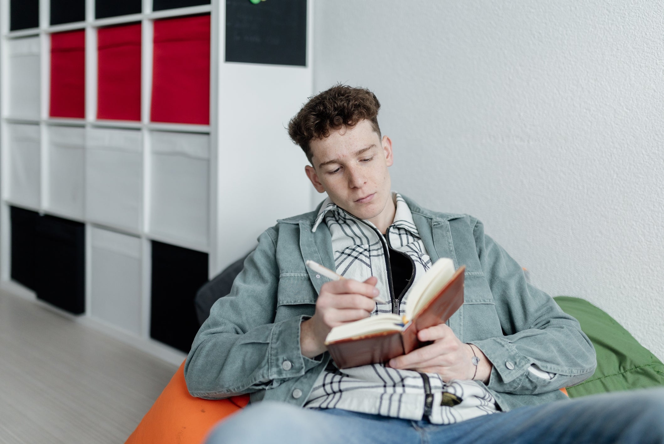 Orang yang duduk di atas beanbag, menulis di buku catatan, mengenakan kemeja kotak-kotak di bawah jaket