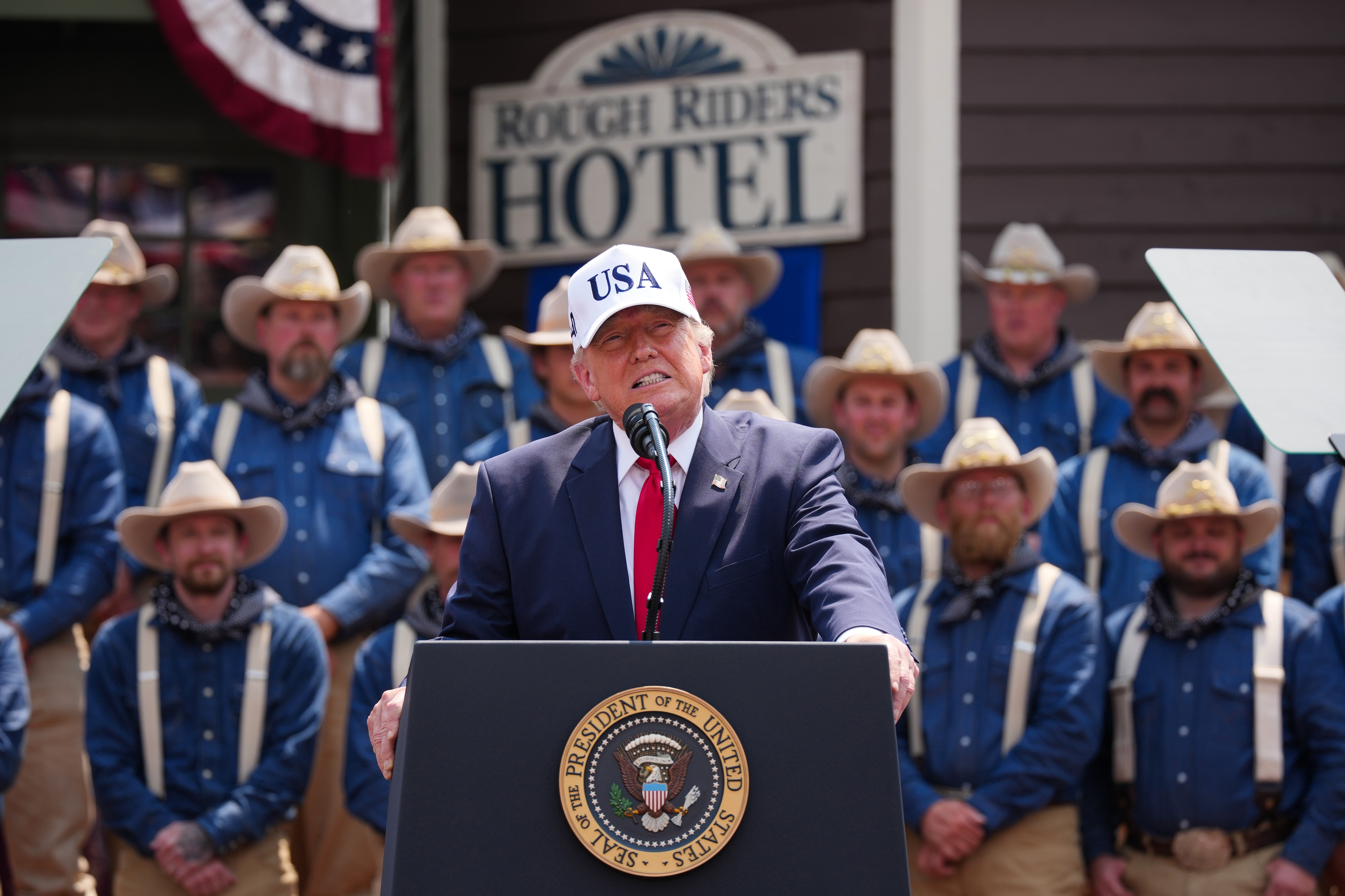 Donald Trump successful a "USA" headdress speaks astatine a podium with a "Rough Riders Hotel" motion behind, surrounded by radical successful occidental attire