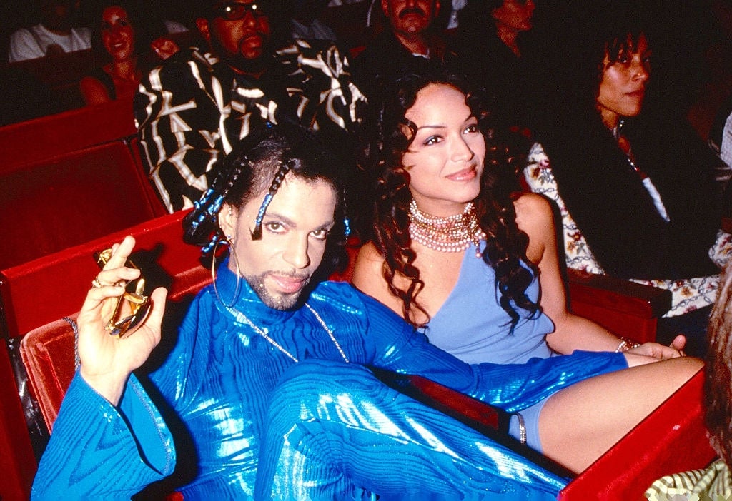 Prince, in an elaborate, shiny outfit, was seated at an event with his hand on Mayte Garcia's lap next to him