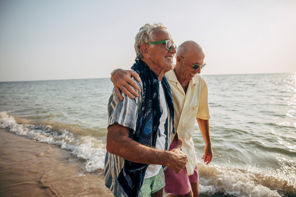 Two older men are happily walking arm-in-arm along a beach, dressed in casual summer shirts and shorts