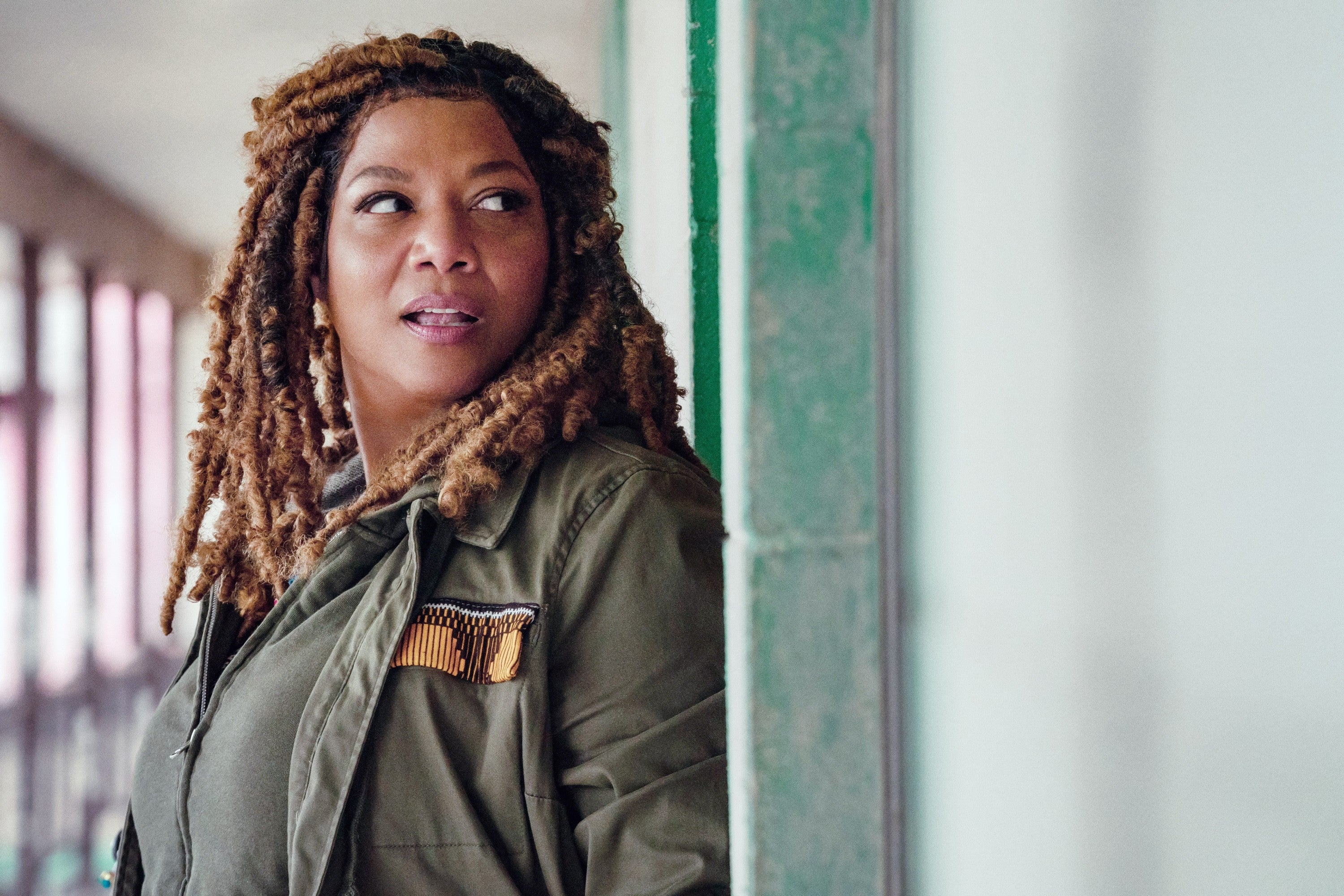 Queen Latifah with locs looks sideways while standing, wearing a jacket with a patch. Background features a corridor