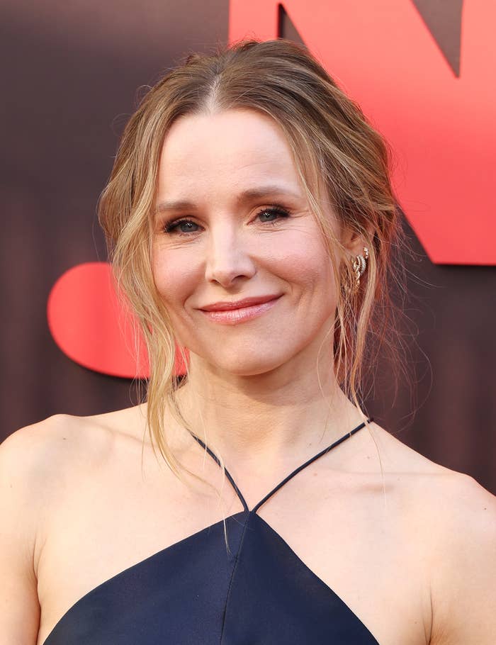 Person on red carpet with a neutral smile, wearing a sleeveless, elegant halter dress. Hair is styled in loose waves