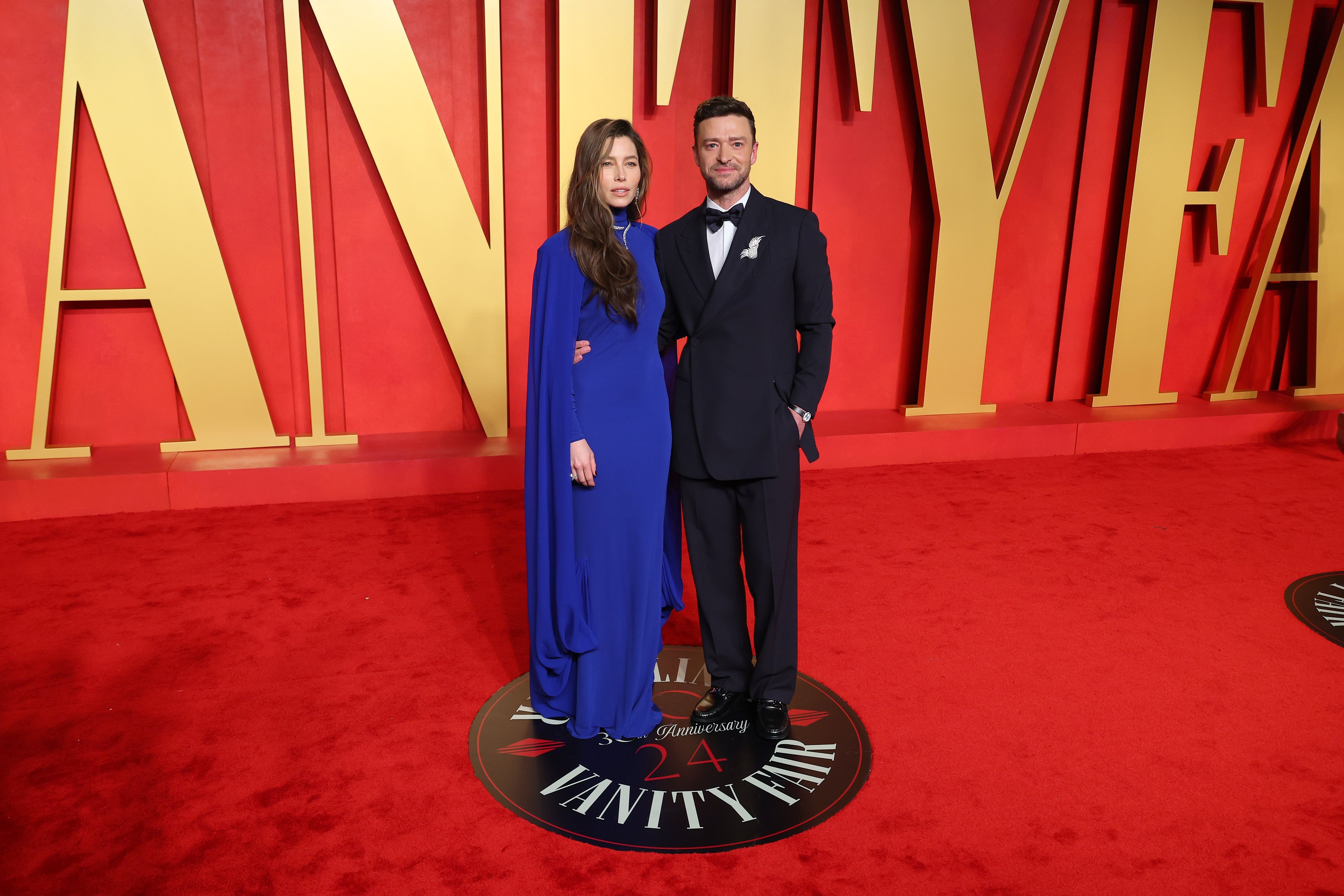 Two people pose on a red carpet. The woman wears a flowing gown, and the man wears a classic tuxedo