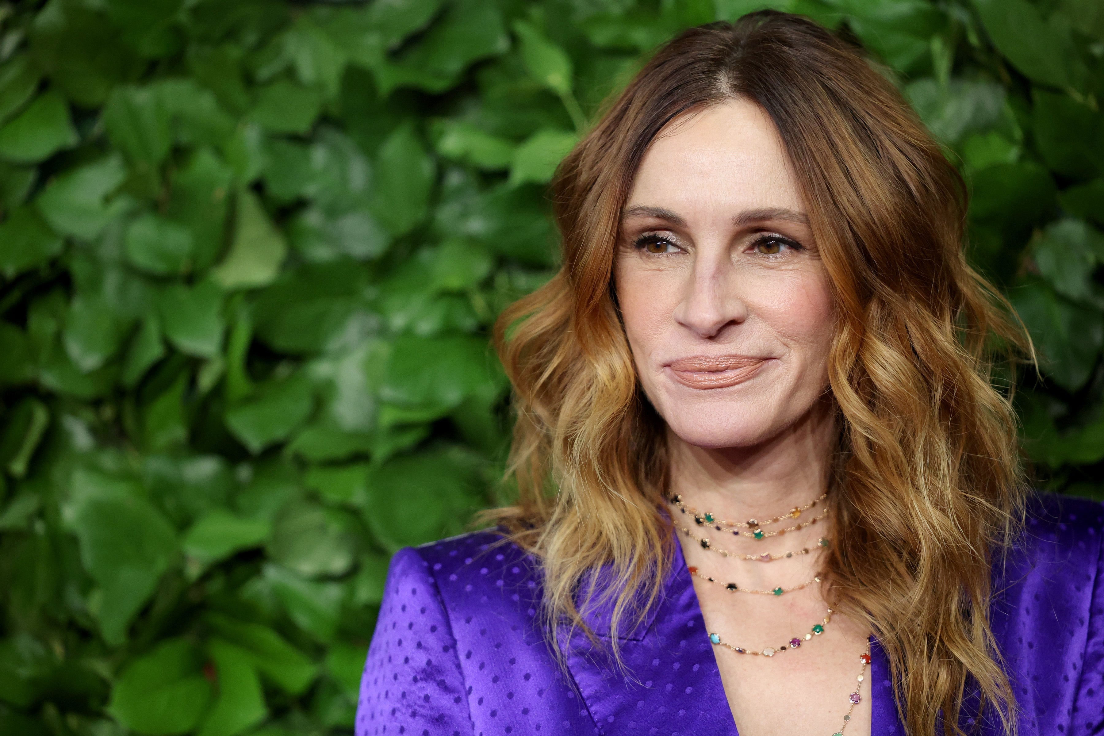 Person with wavy hair wearing a patterned suit jacket and layered necklaces, standing in front of a leafy background