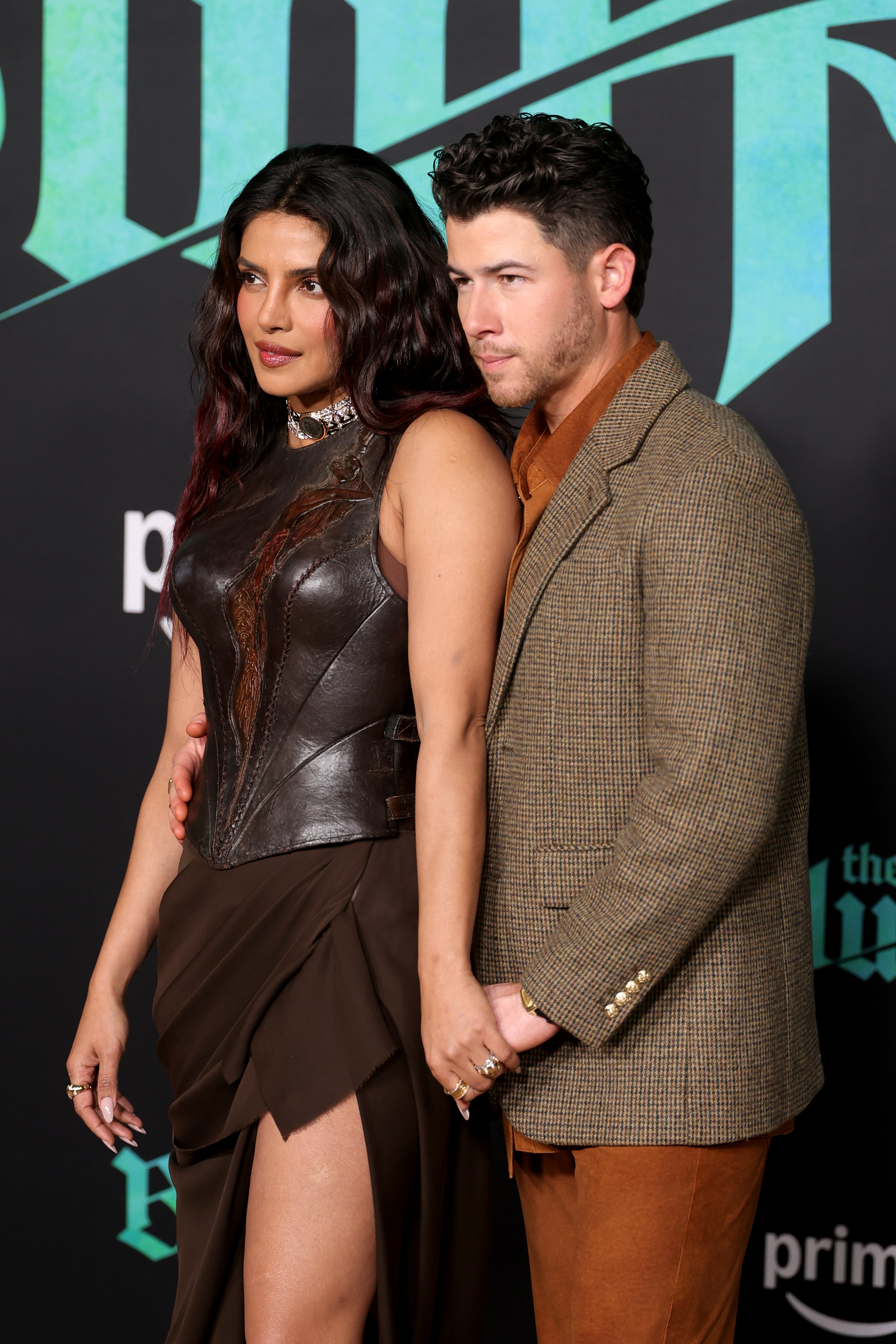 A couple poses on the red carpet. One is wearing a leather top and skirt, while the other is in a textured blazer and pants