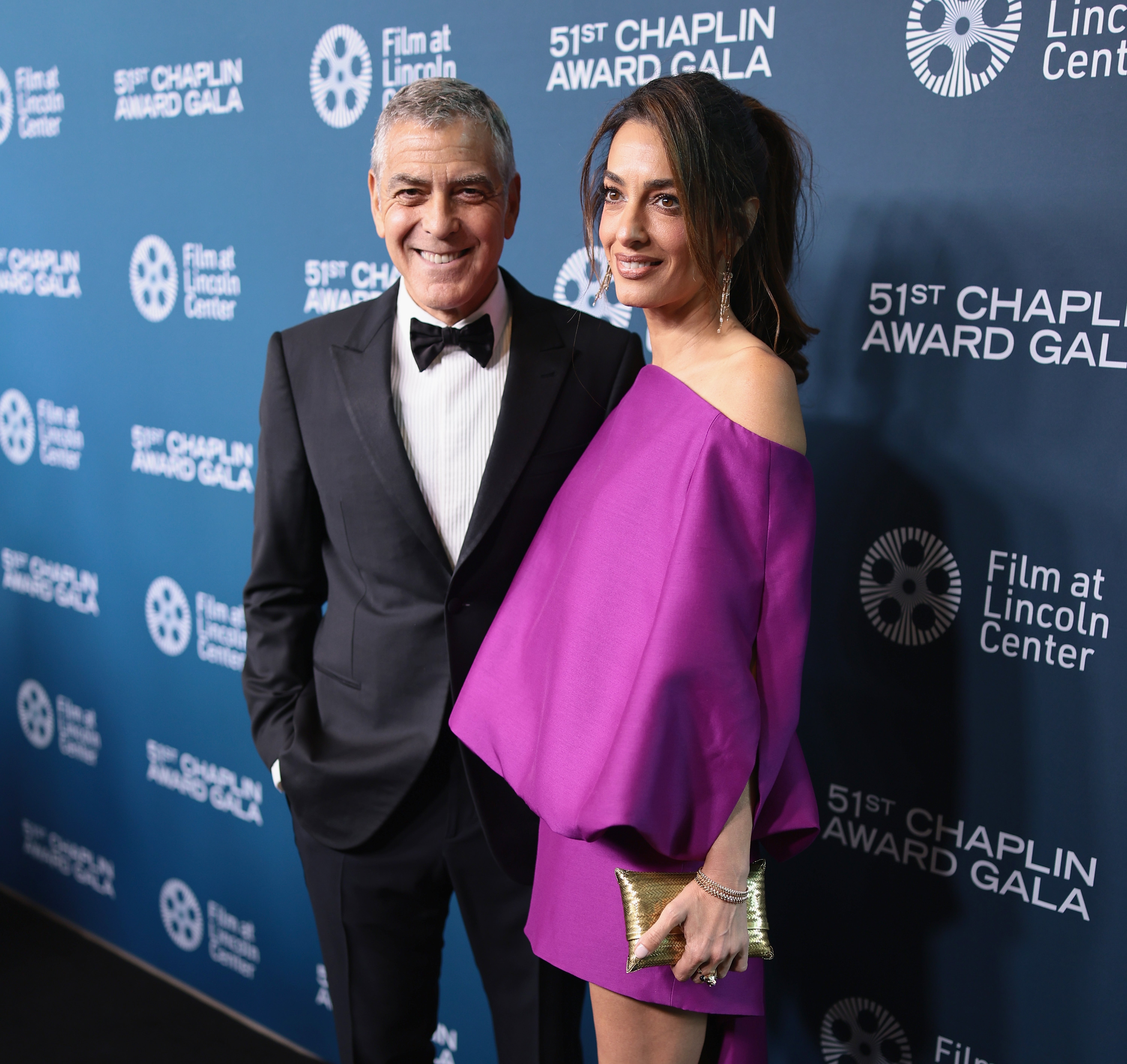 A couple poses on a red carpet at an award gala. The man wears a tuxedo; the woman wears an off-the-shoulder, draped dress