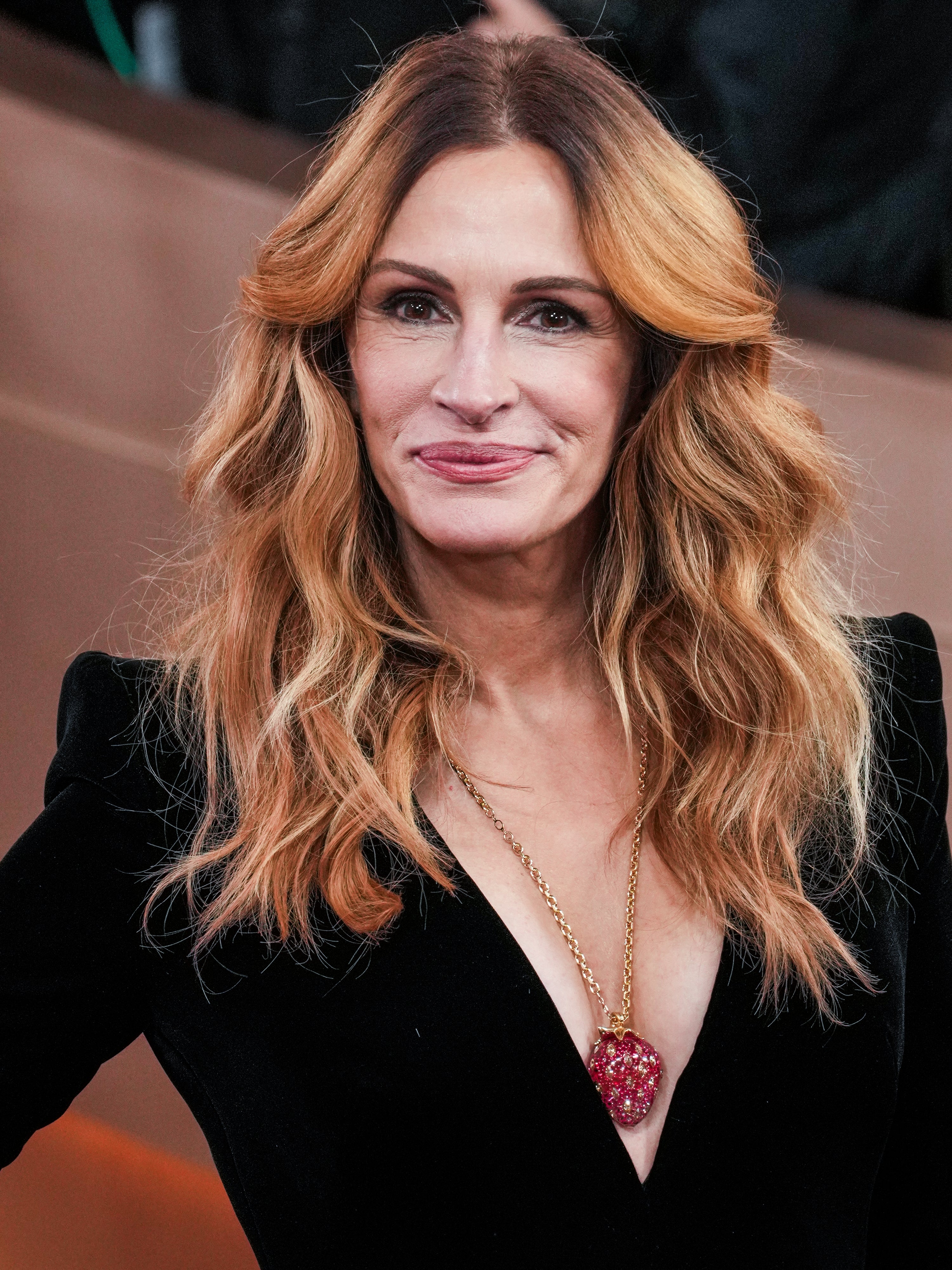 A person wearing a black velvet outfit with a deep neckline and a long pendant necklace, smiling and looking toward the camera