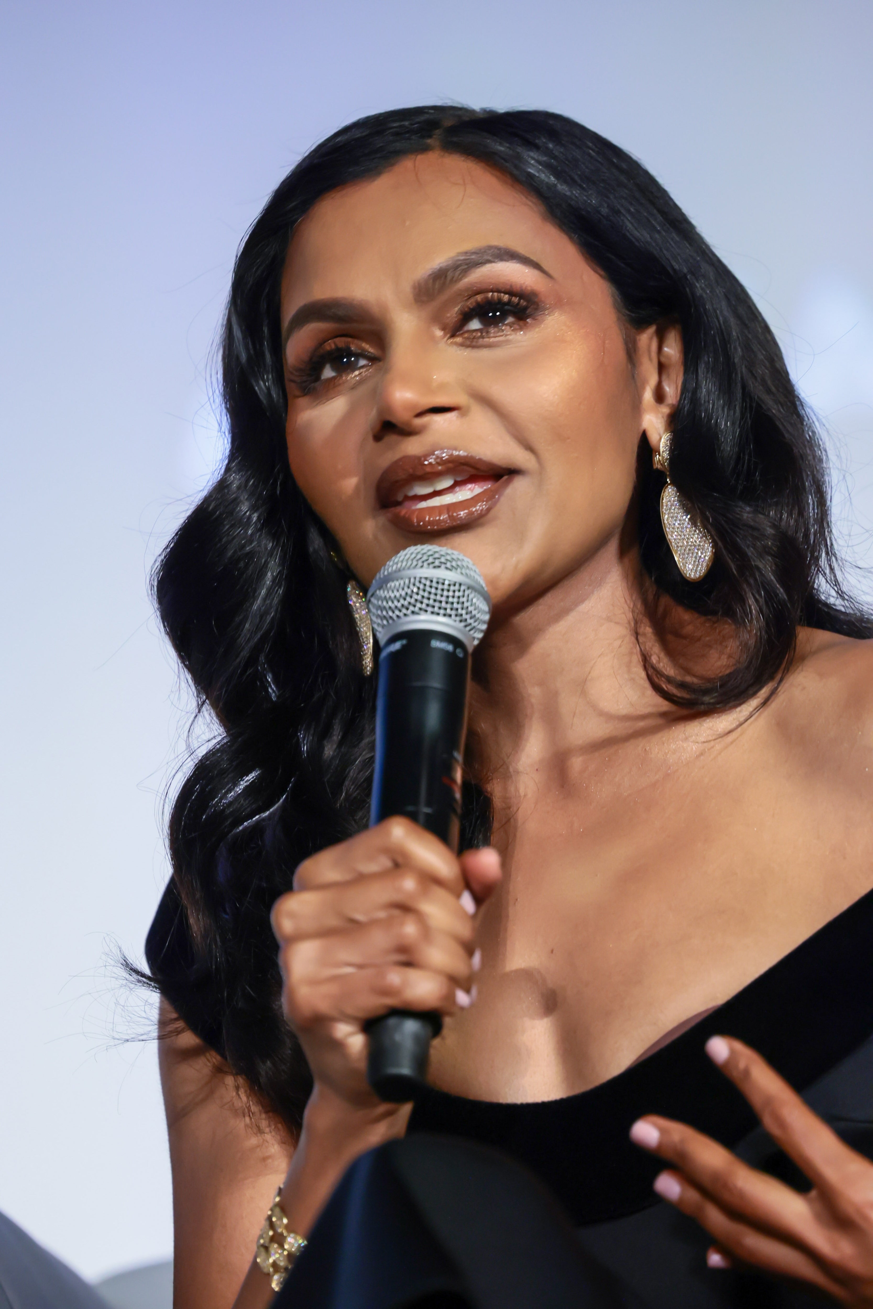 Person speaking into a microphone, wearing an elegant, off-shoulder dress and large earrings