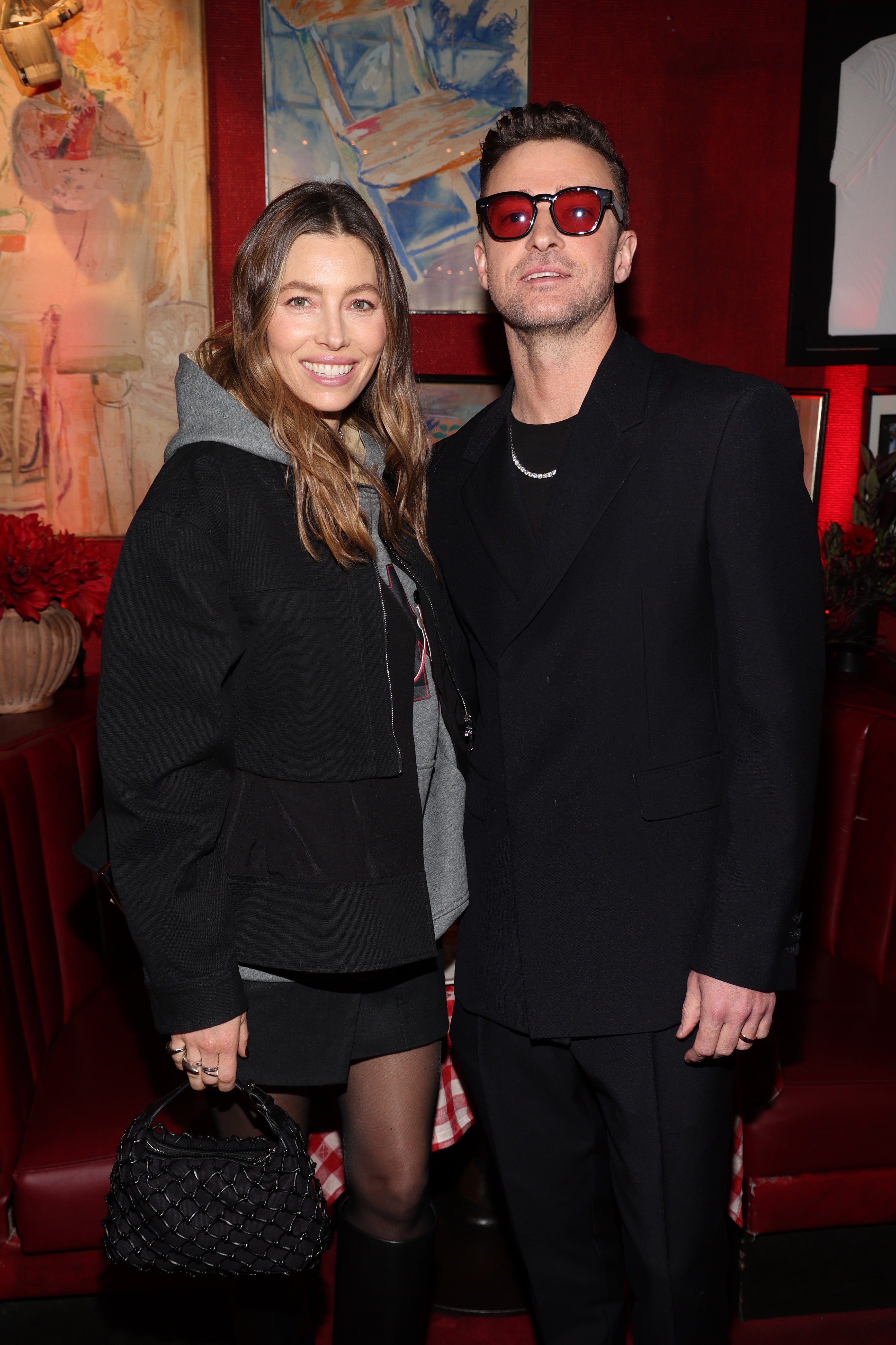 Two people posing together indoors; one in a hoodie and jacket, the other in a suit with sunglasses