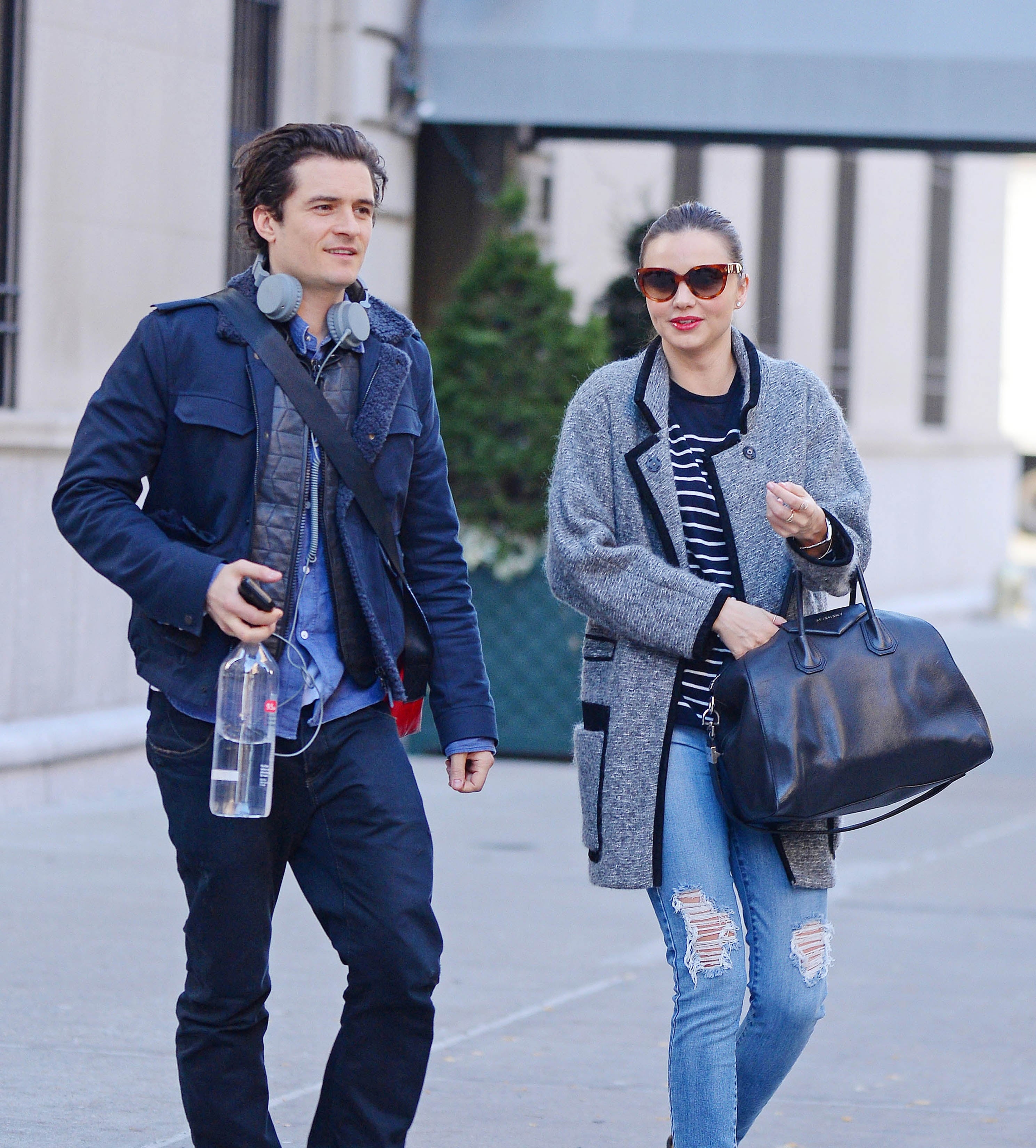 Two people walking on a city street. One is wearing a jacket with headphones around their neck, and the other in a long coat and sunglasses, holding a handbag