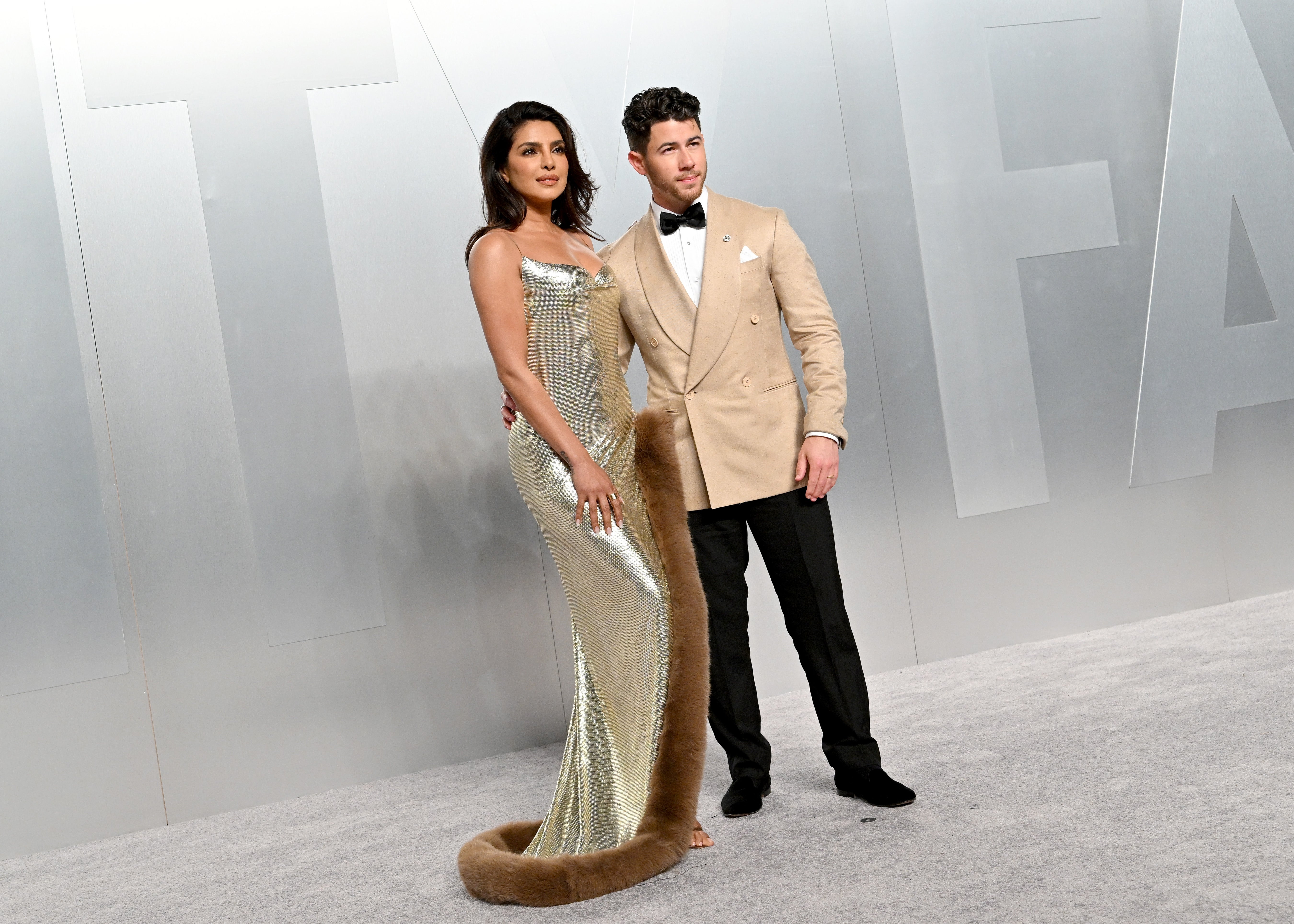 A couple on a red carpet; the woman in a metallic gown with a fur trim, the man in a tailored beige suit with a bow tie