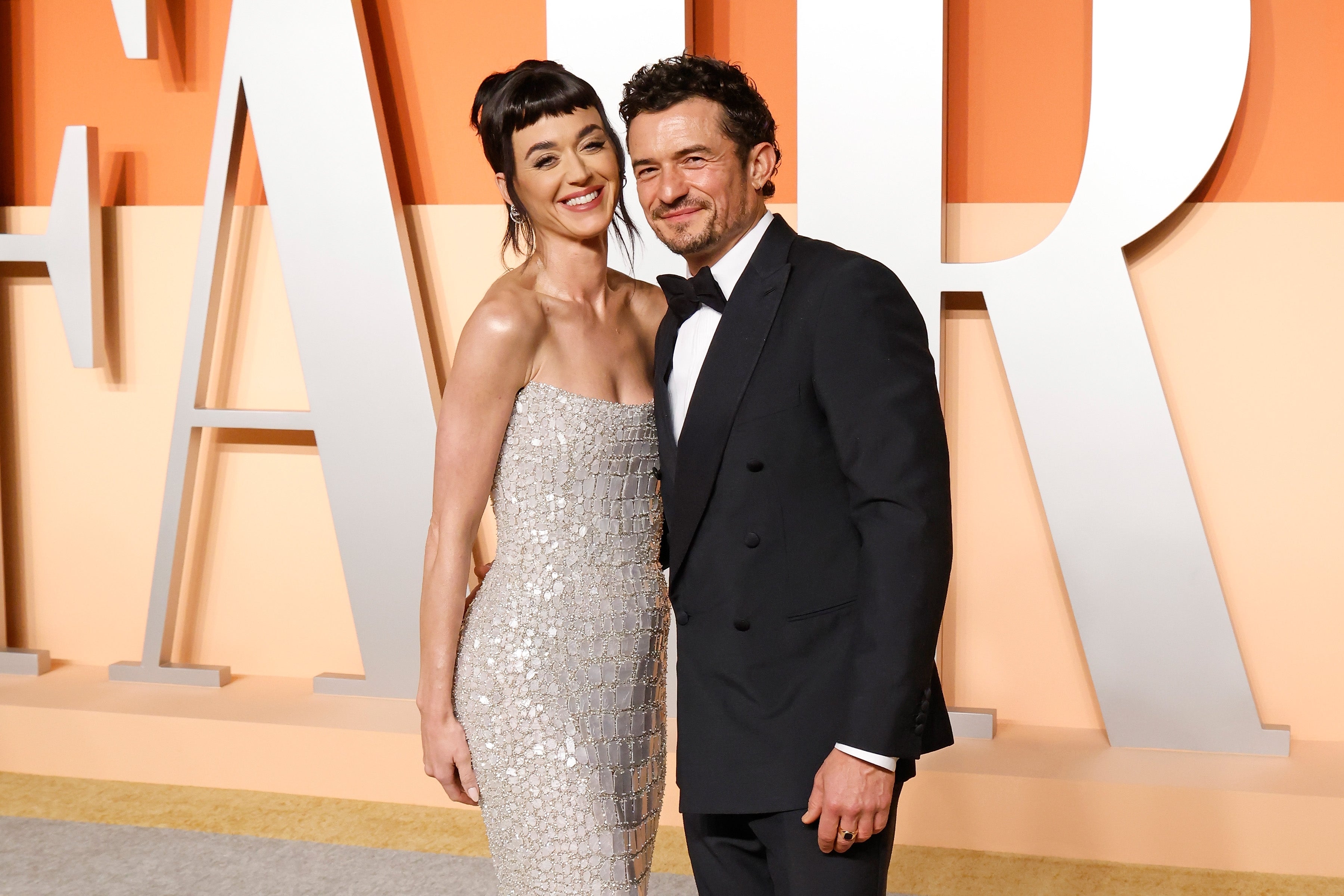 A couple poses on a red carpet; the woman wears a strapless, sequined dress, and the man is in a classic tuxedo