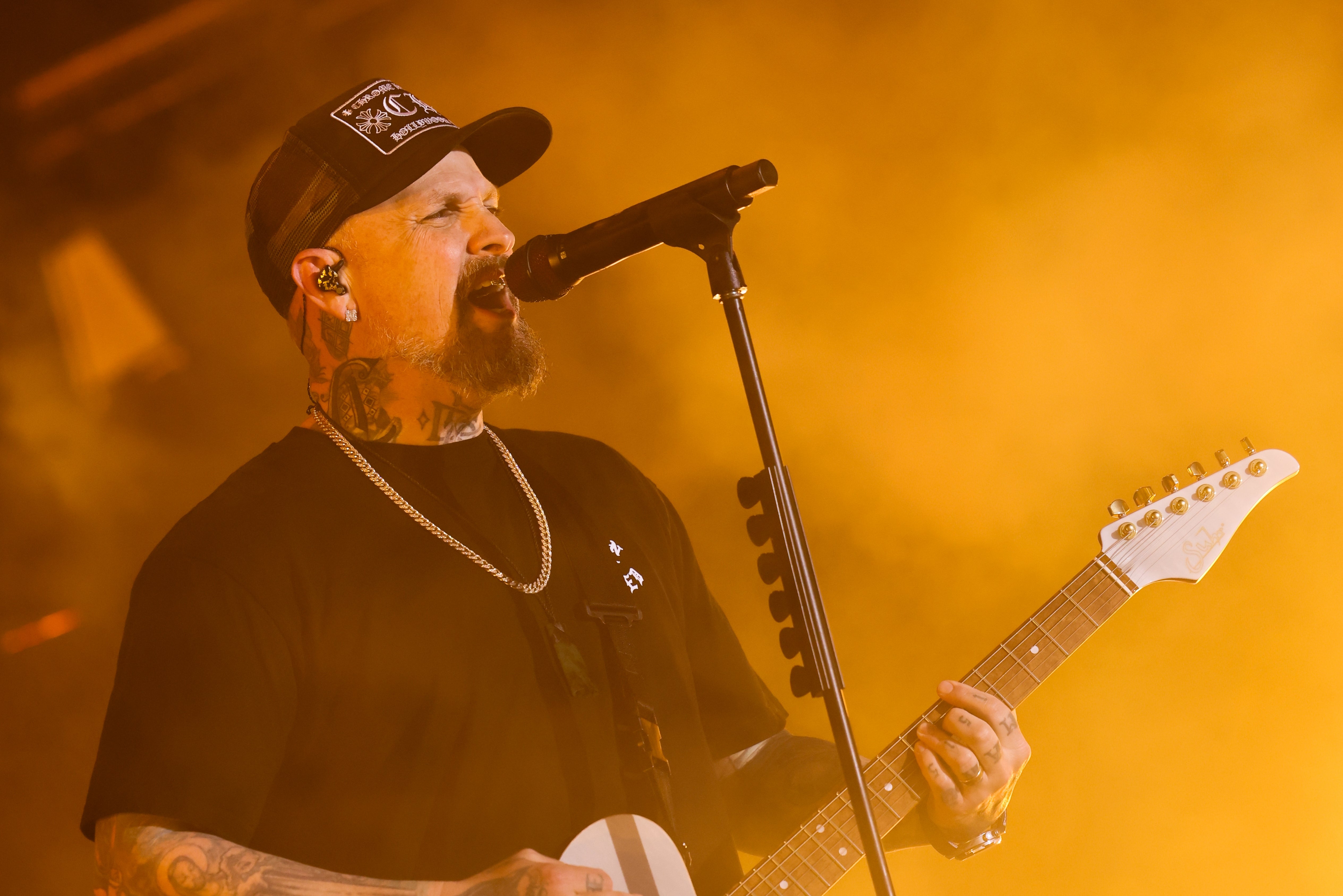 Musician performing energetically on stage, singing into a microphone and playing guitar. Wearing a cap, chain necklace, and tattoos visible