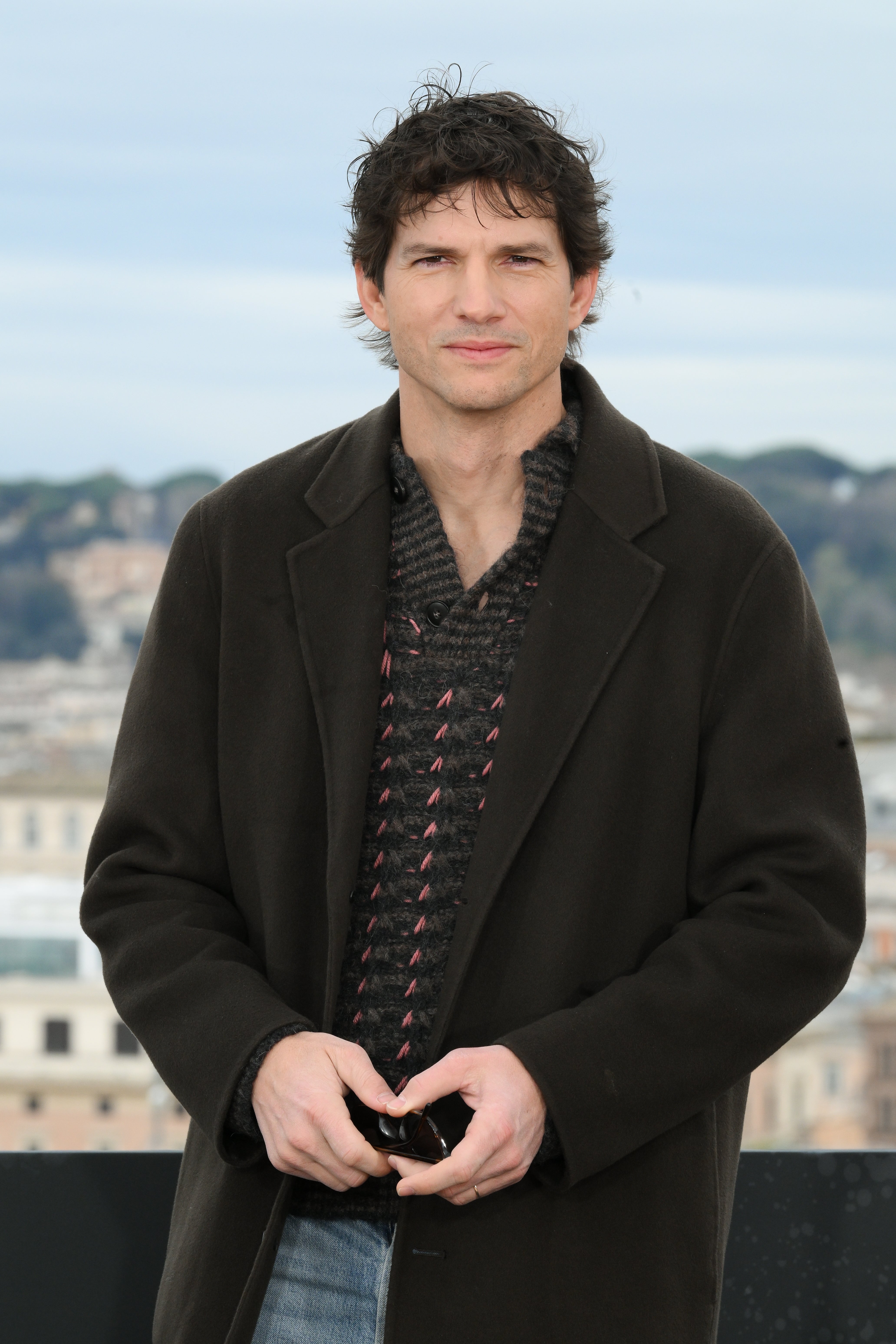 A person stands outside in a coat and patterned sweater, hands together, with a blurred cityscape in the background