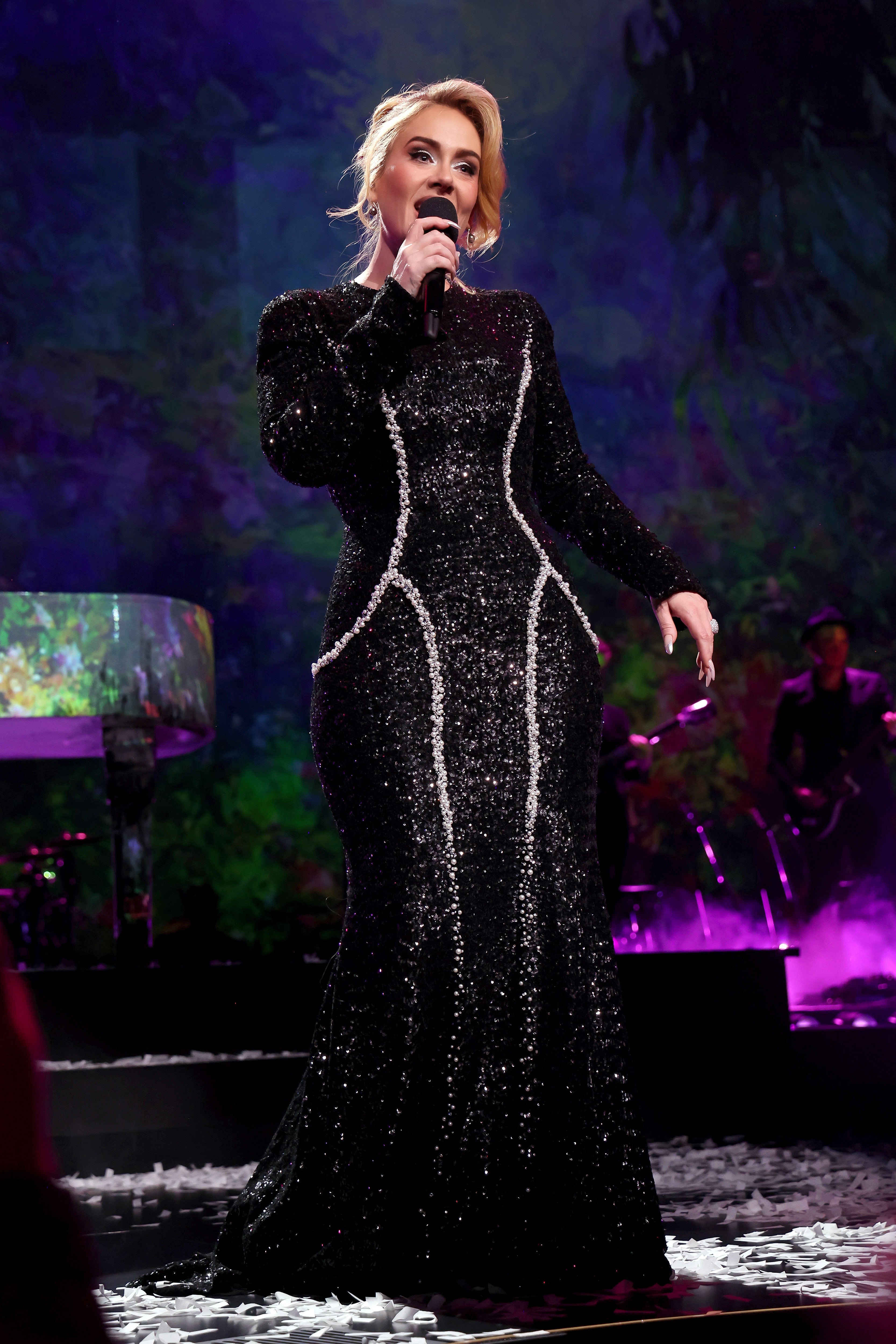 Person in a glittering, form-fitting black gown with silver accents sings on stage with band and piano in the background