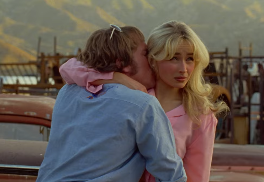 A couple embraces outdoors. The woman, in a stylish outfit, looks ahead while the man whispers closely to her. Desert landscape in the background