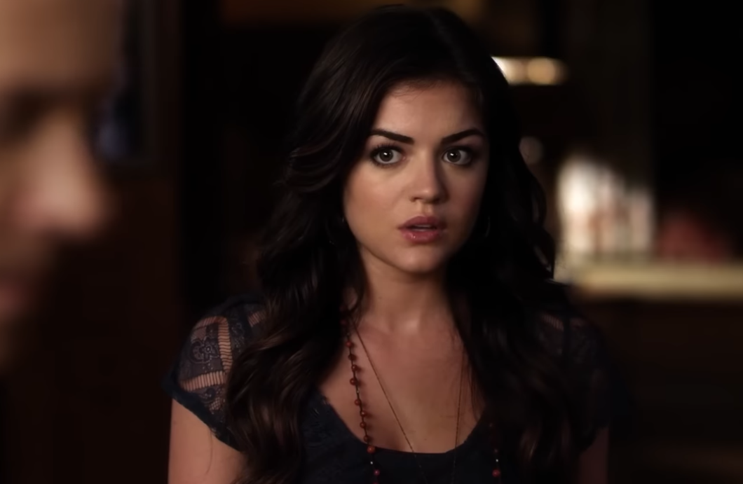 A woman with long dark hair and a surprised expression stands indoors, wearing a lace top and a beaded necklace