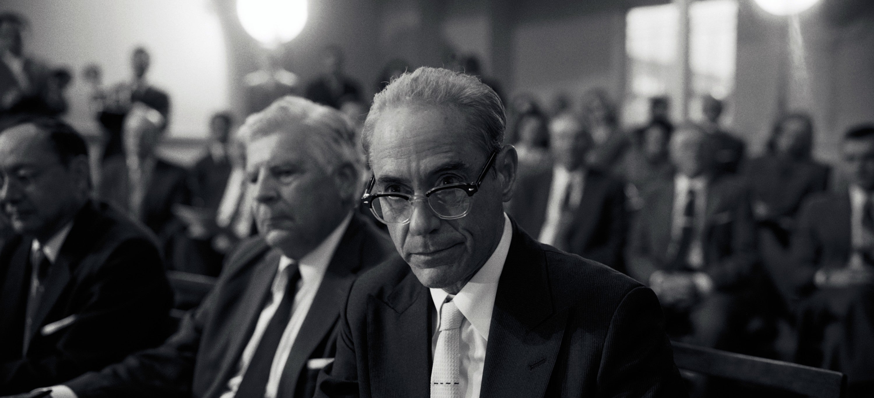 Robert Disney Jr in a suit and glasses sits attentively among a group of formally dressed individuals at an indoor event