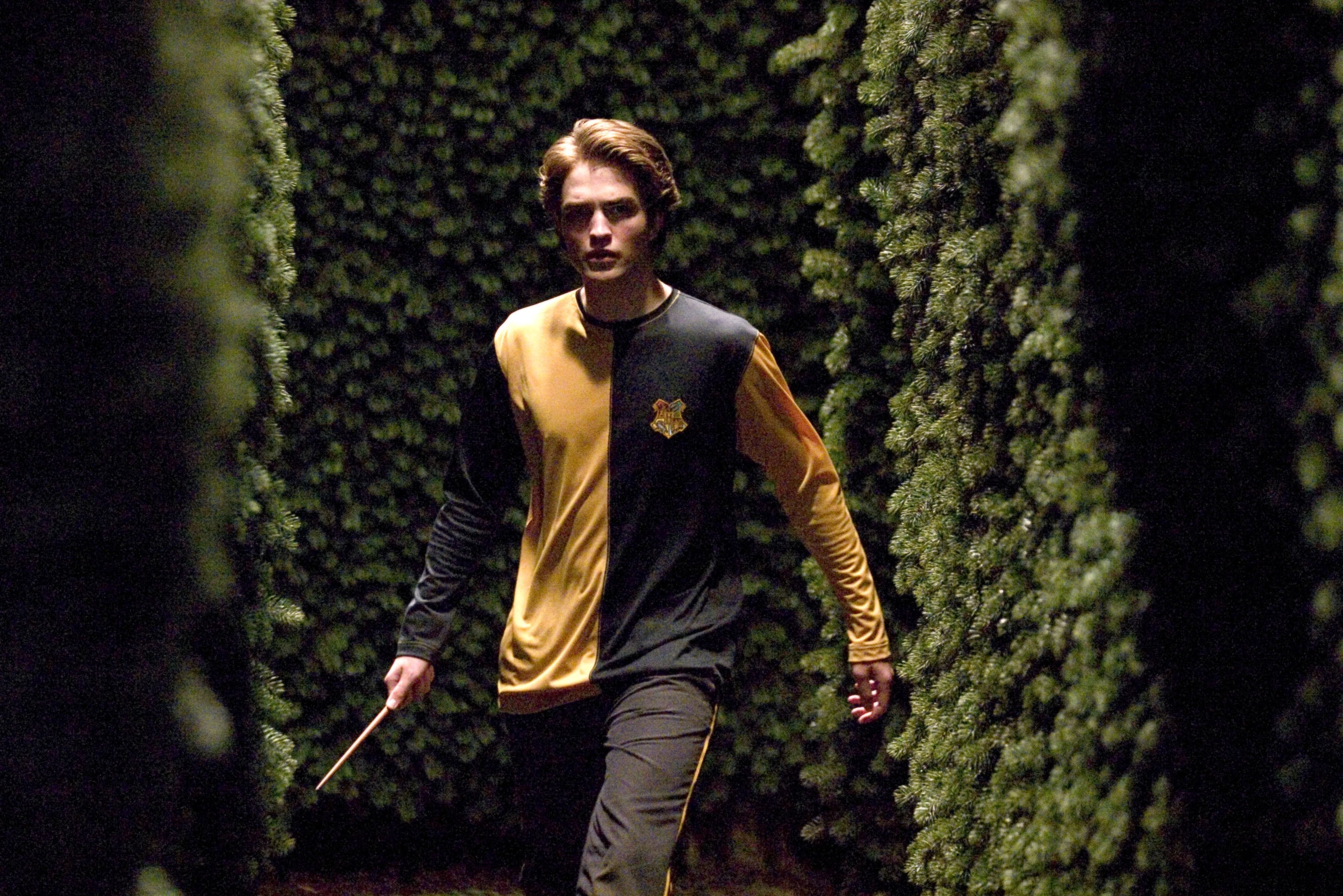 Robert Pattinson walking through a hedge maze wearing a two-toned athletic outfit, holding a wand, appearing focused and determined