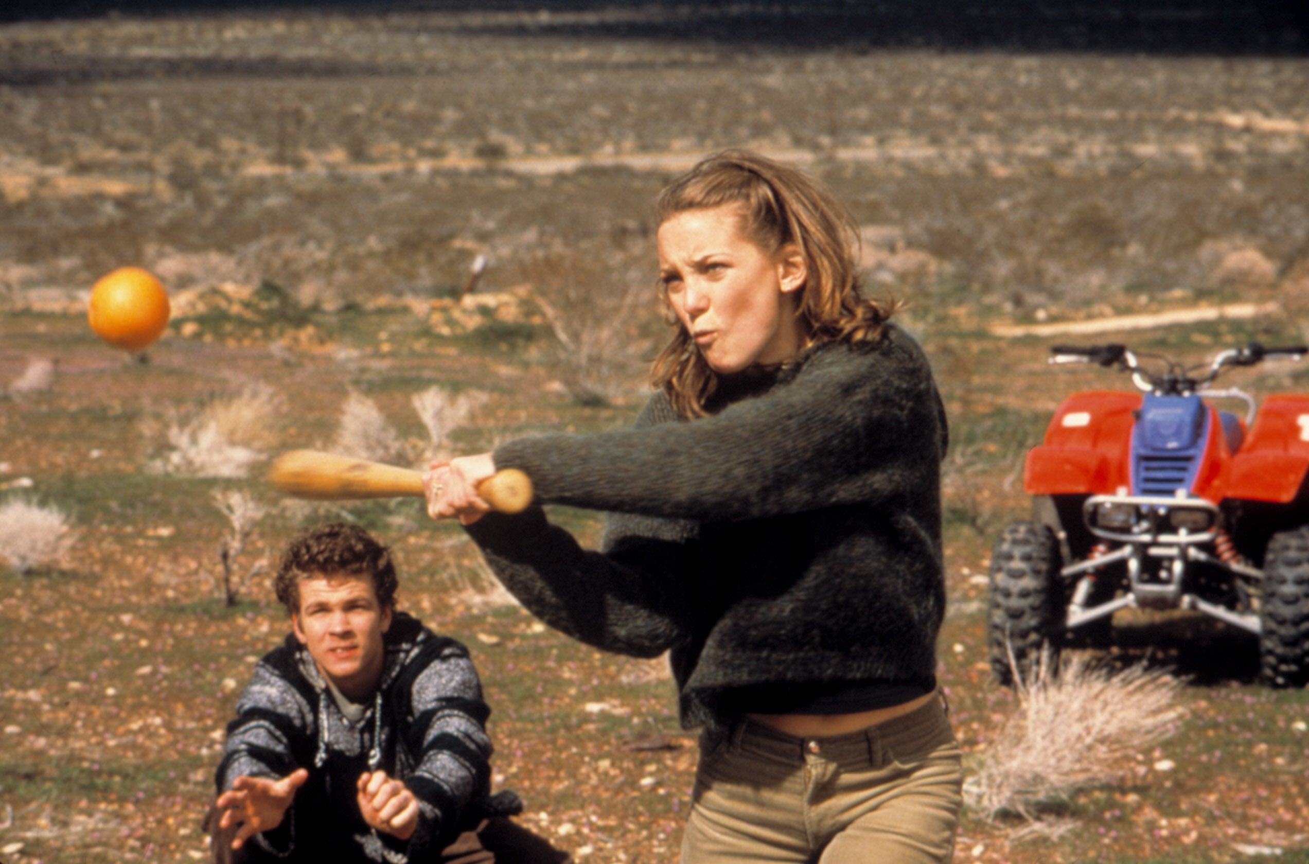 Younger Kate Hudson swinging a wooden bat at an orange in a desert setting, with another person watching. An ATV is parked nearby