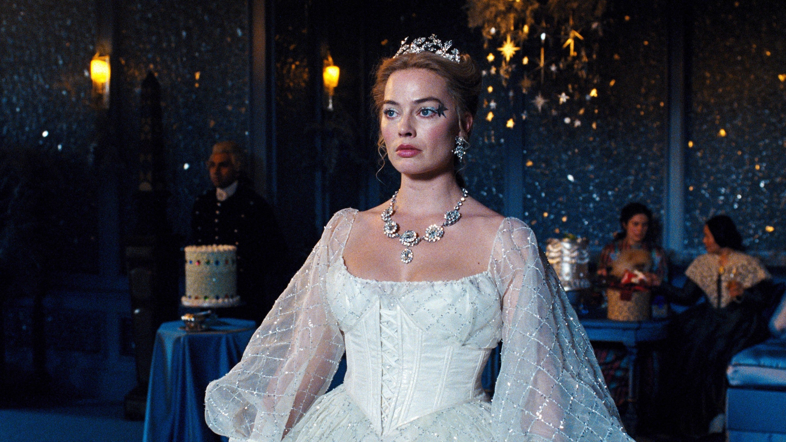 Margot Robbie in an elegant gown and ornate jewelry stands in a formal event setting, surrounded by guests and a decorative cake in the background