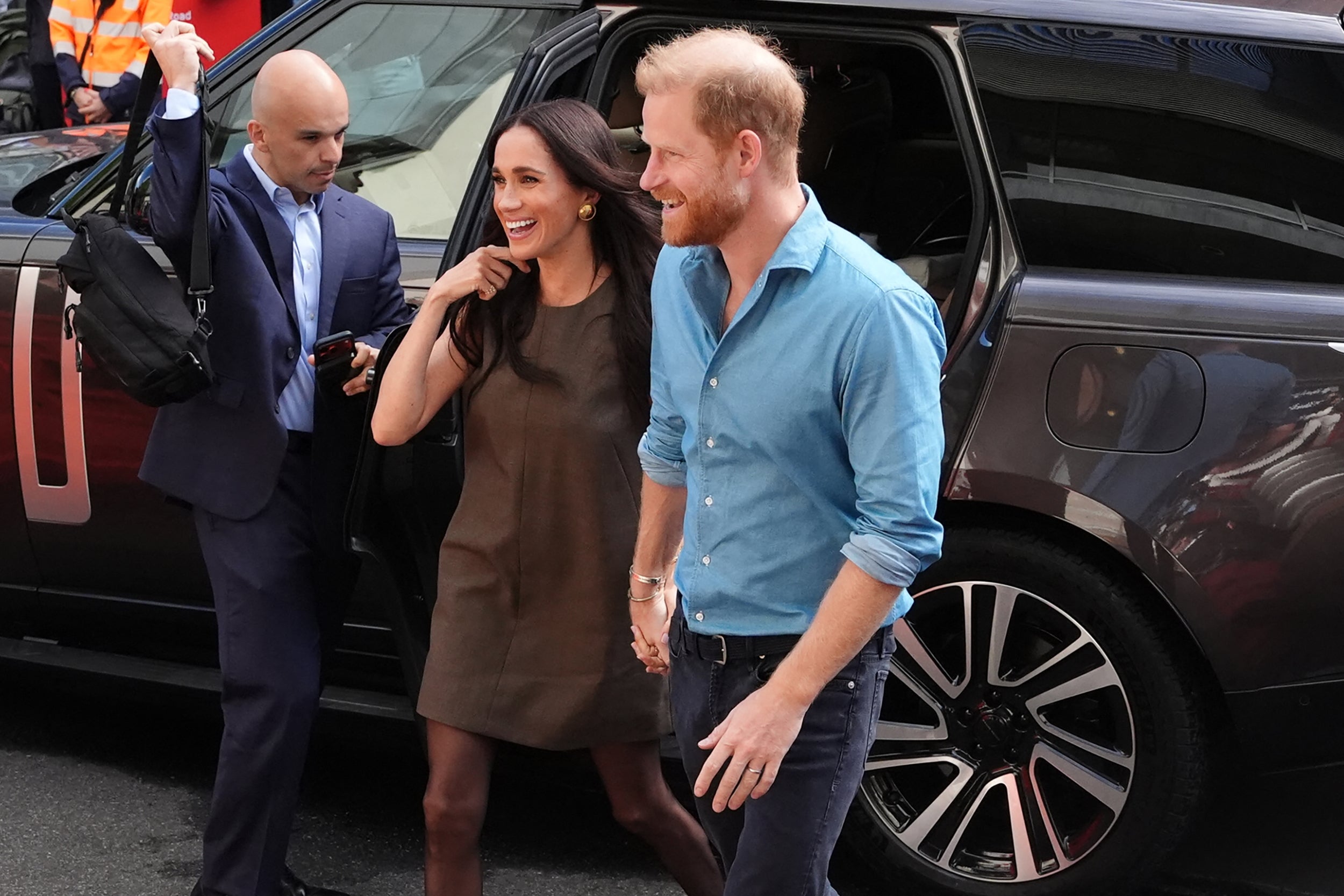 Two people smiling, exiting a car. One is in a casual blue shirt, the other in a simple dress, accompanied by an attendant with a bag
