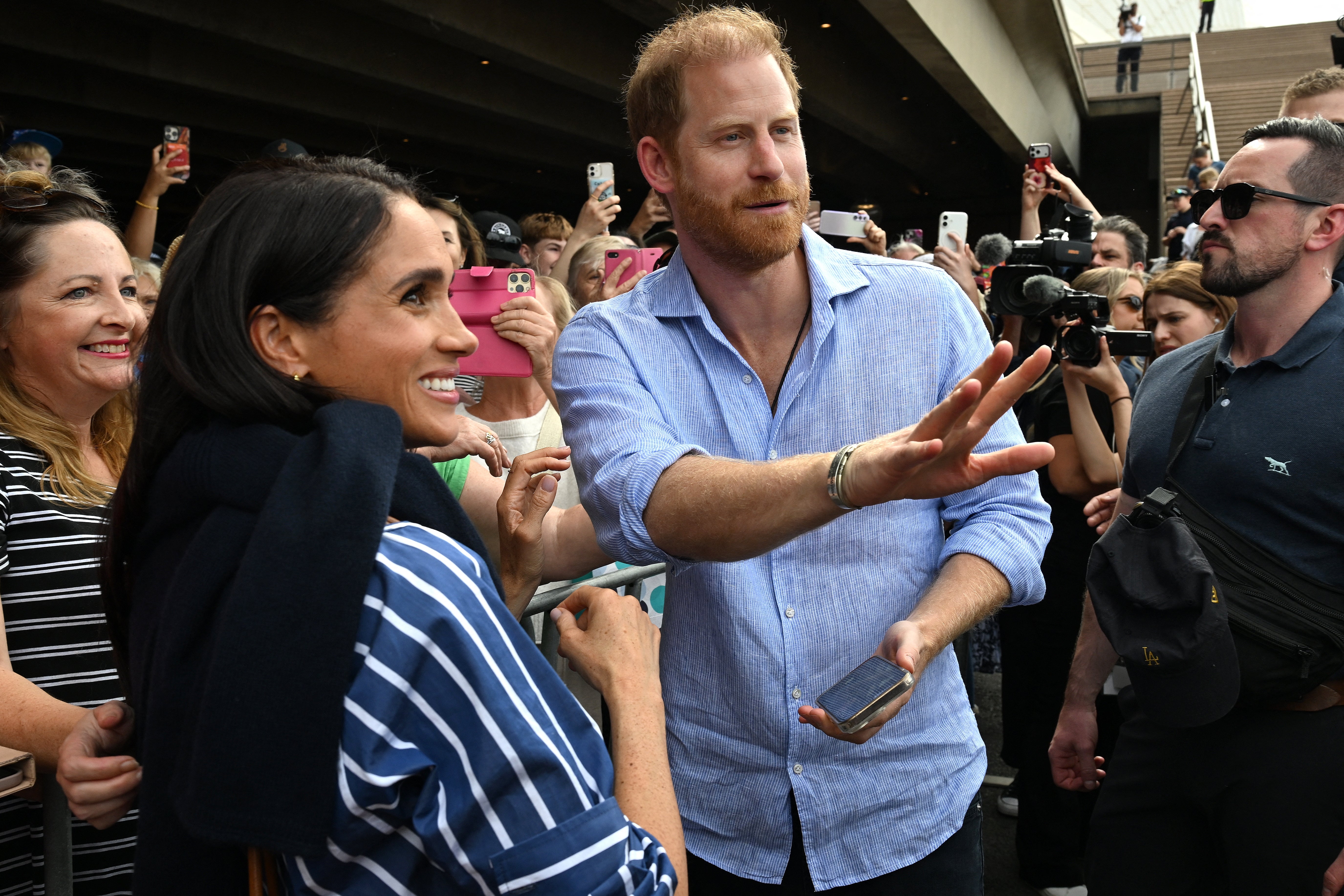 A man and woman interact with a crowd, smiling and waving. The woman wears a striped outfit, and the man is in a casual button-up shirt